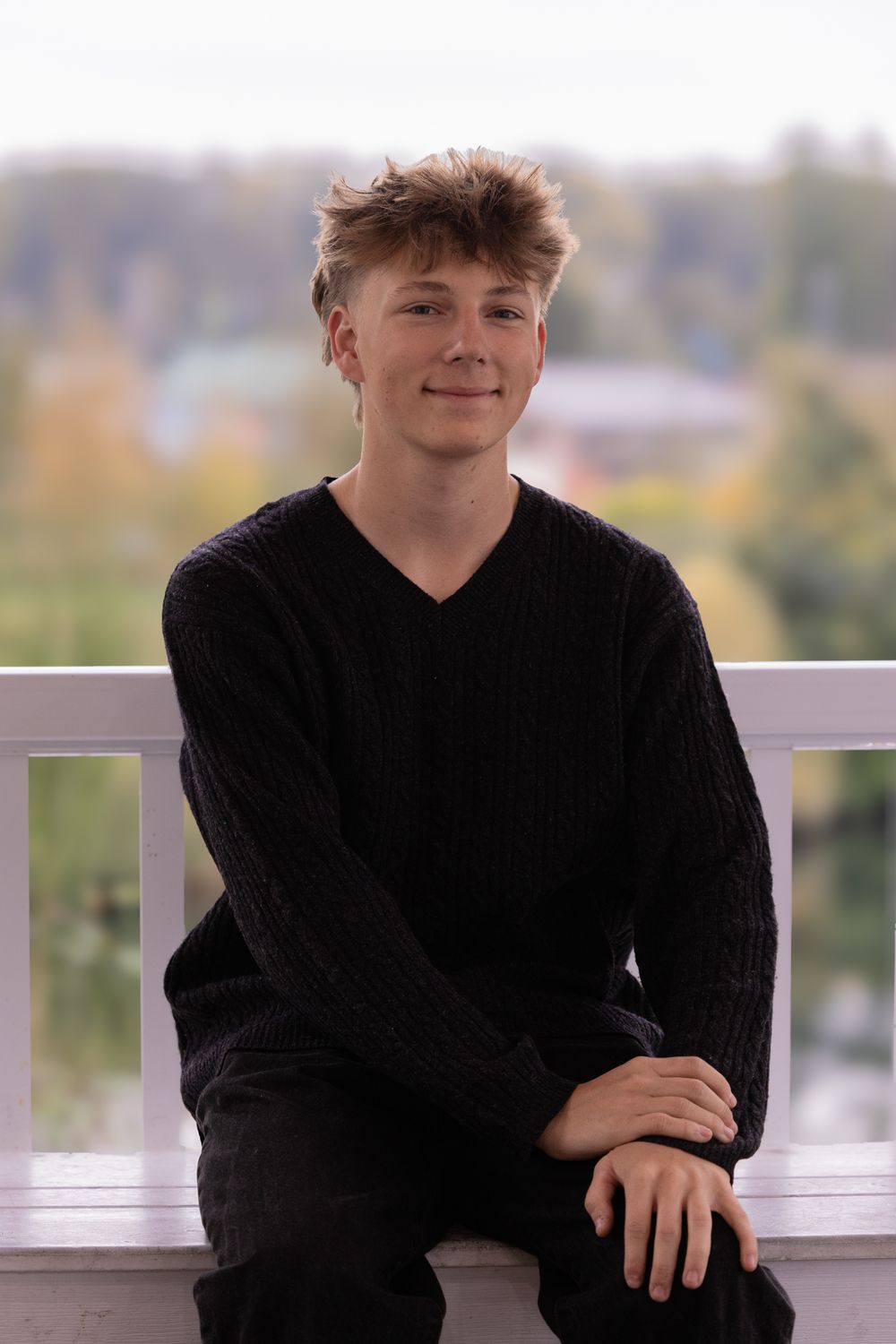 Young man in black cable-knit sweater sitting on white railing, smiling outdoors with blurred autumn background.