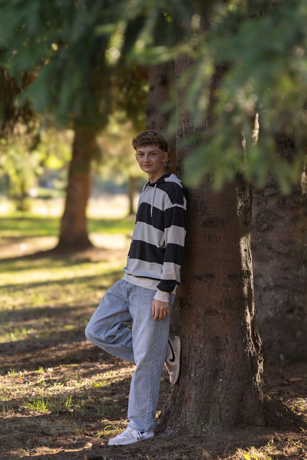 Teen boy in striped hoodie and jeans leaning against a tree trunk in a sunny park setting.
