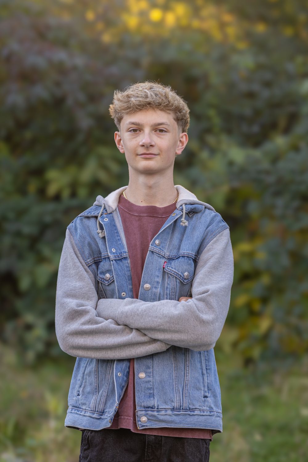 Young man with curly blonde hair in denim jacket stands with arms crossed outdoors among autumn foliage.