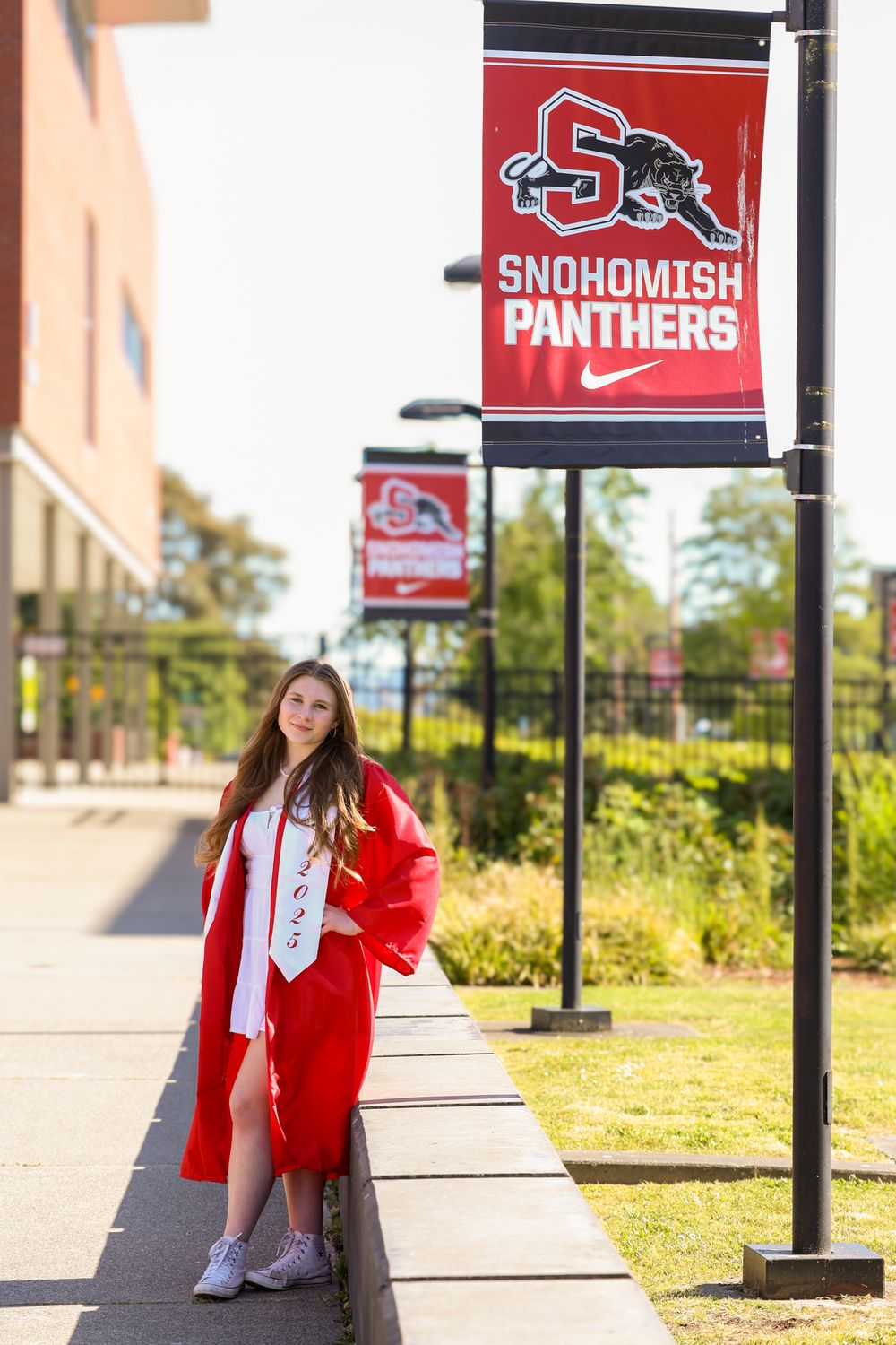 A young woman in red graduation cap and gown poses beside Snohomish Panthers school banners on a sunny campus walkway.