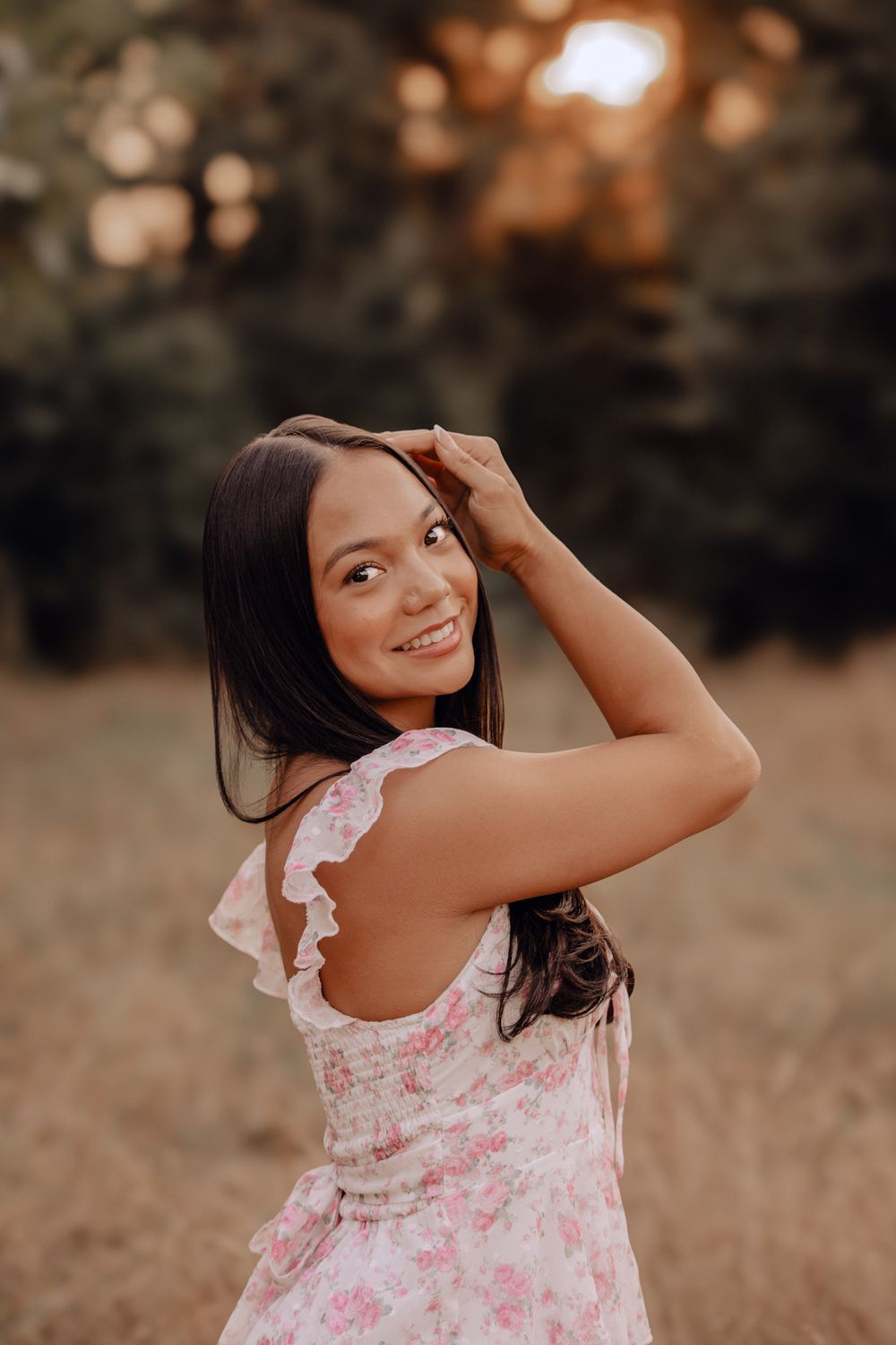 Young woman in floral pink dress smiling over shoulder at sunset in outdoor park setting.