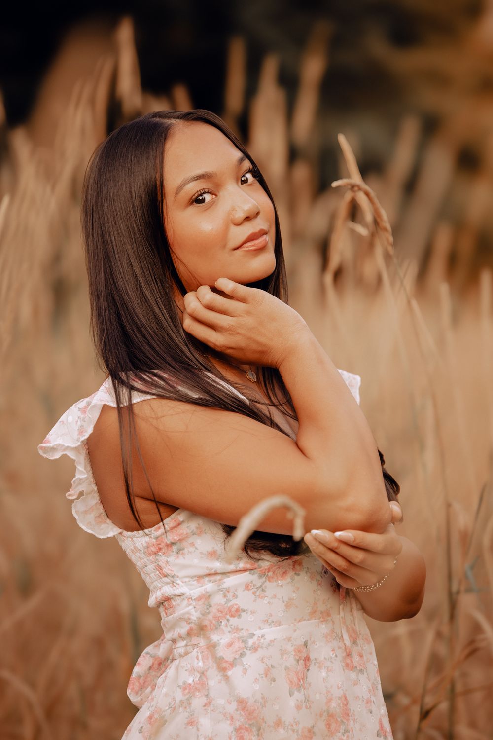 Young woman in floral dress posing gracefully among tall golden grasses in a warm, sunlit outdoor setting.