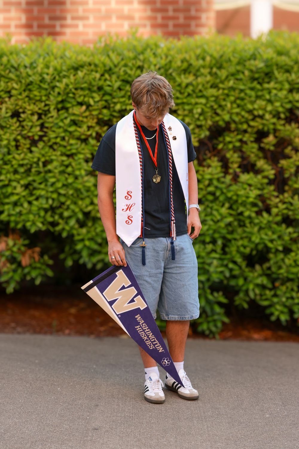 High school graduate holding University of Washington pennant, wearing white SHS stole and medal, standing outdoors.