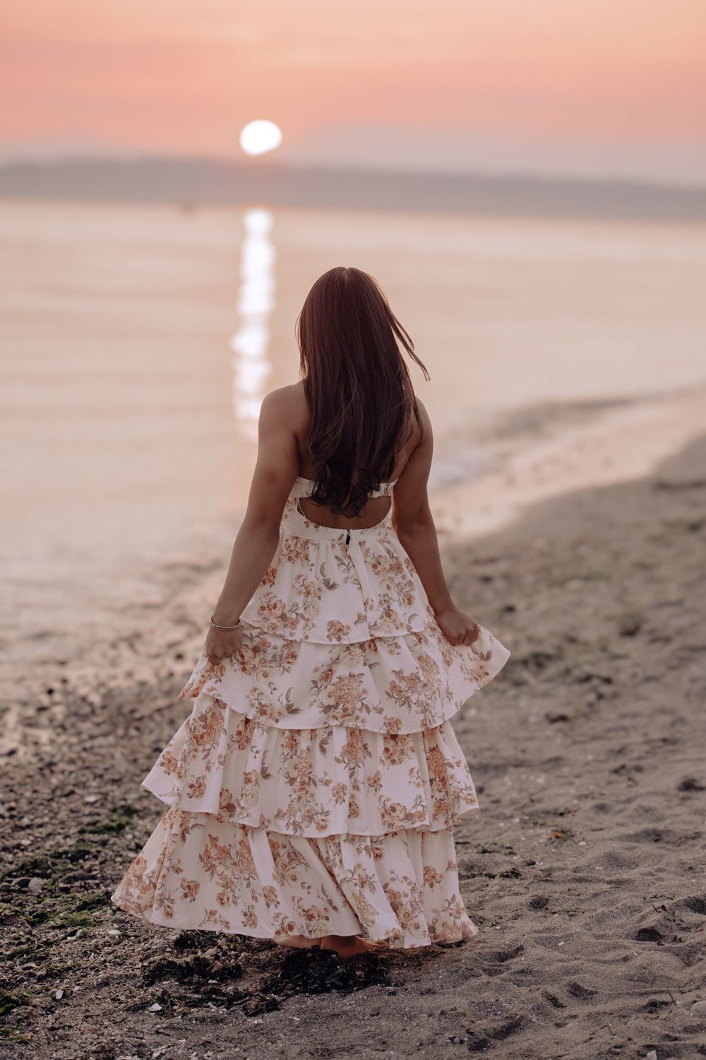 Woman in floral tiered maxi dress standing on beach at sunset, viewed from behind with long hair flowing.