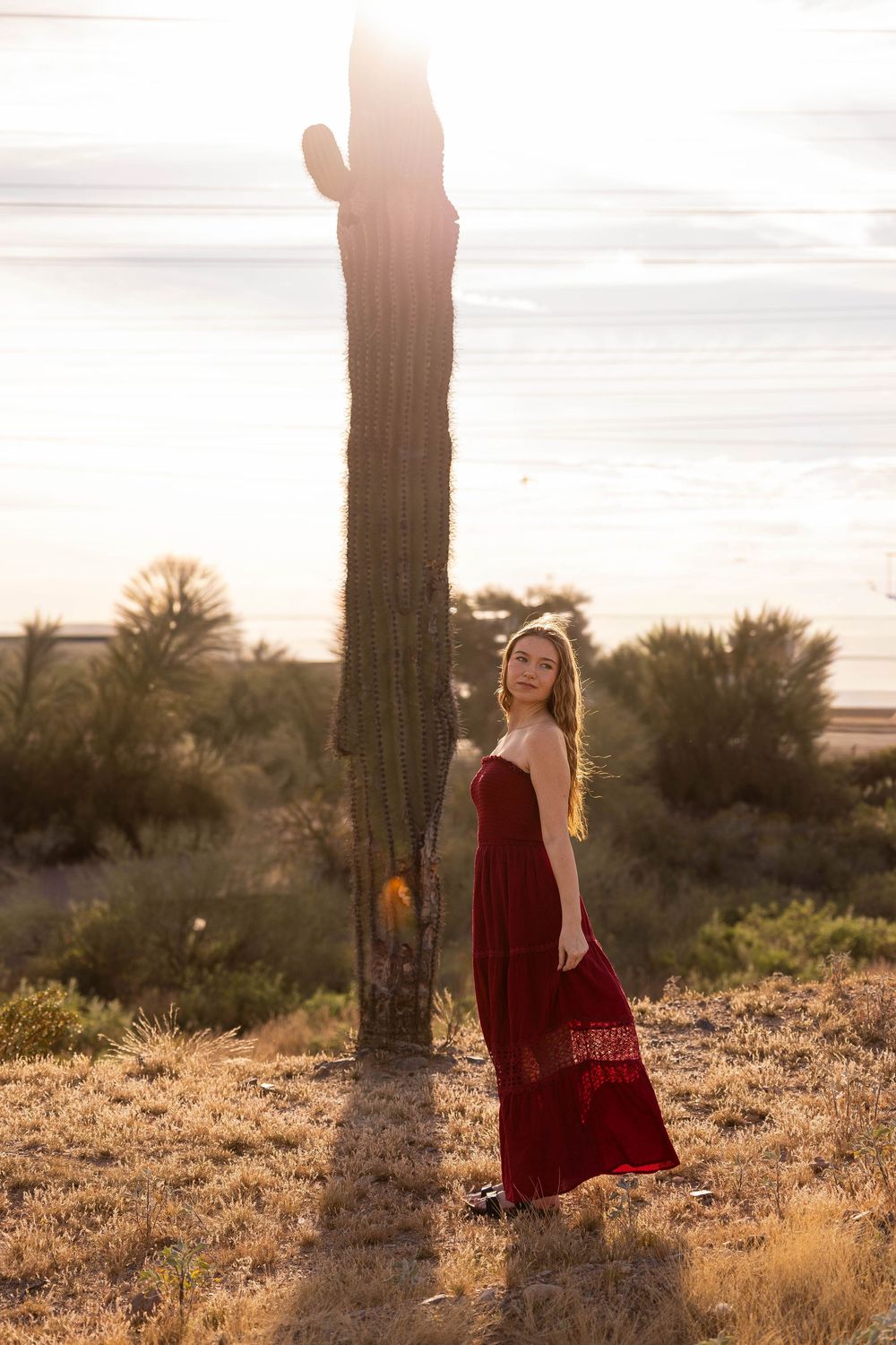 Young woman in red dress poses beside tall saguaro cactus at golden hour sunset in Arizona desert.