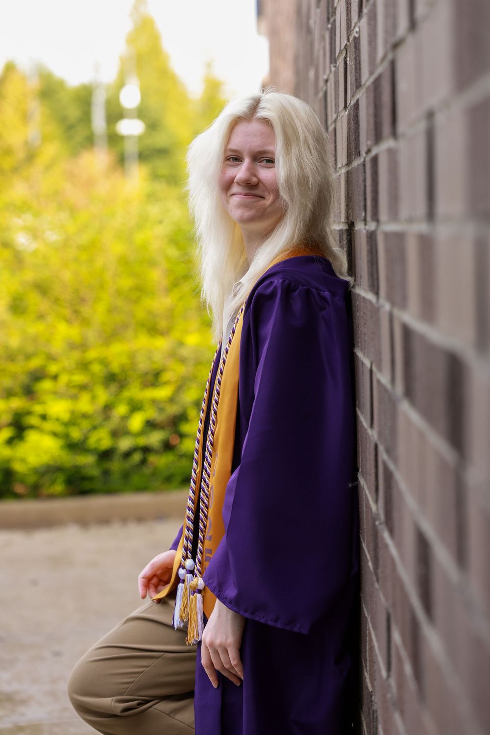 Smiling blonde graduate in purple cap and gown with gold honor cords leans against brick wall outdoors.