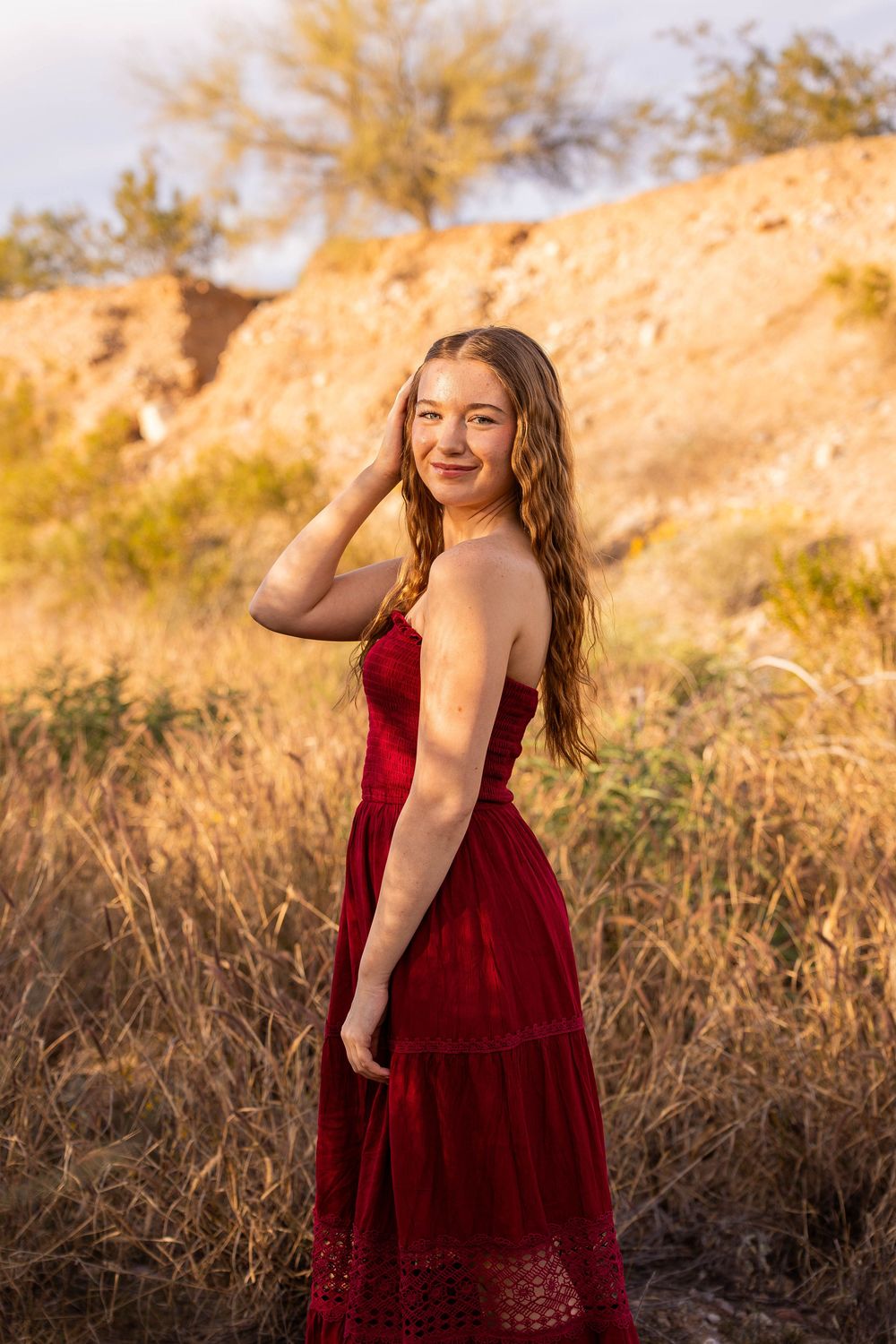 Young woman in red dress poses gracefully in golden field at sunset with rocky hillside backdrop.
