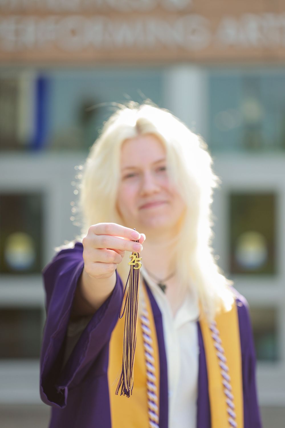 Blonde woman in purple graduation gown holds tassel toward camera outside Performing Arts building.