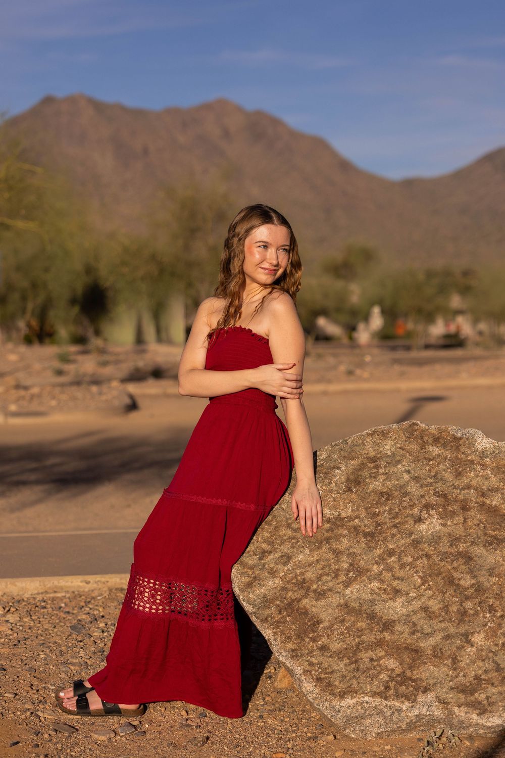 Young woman in red maxi dress posing by a boulder in Arizona desert landscape with mountain backdrop.