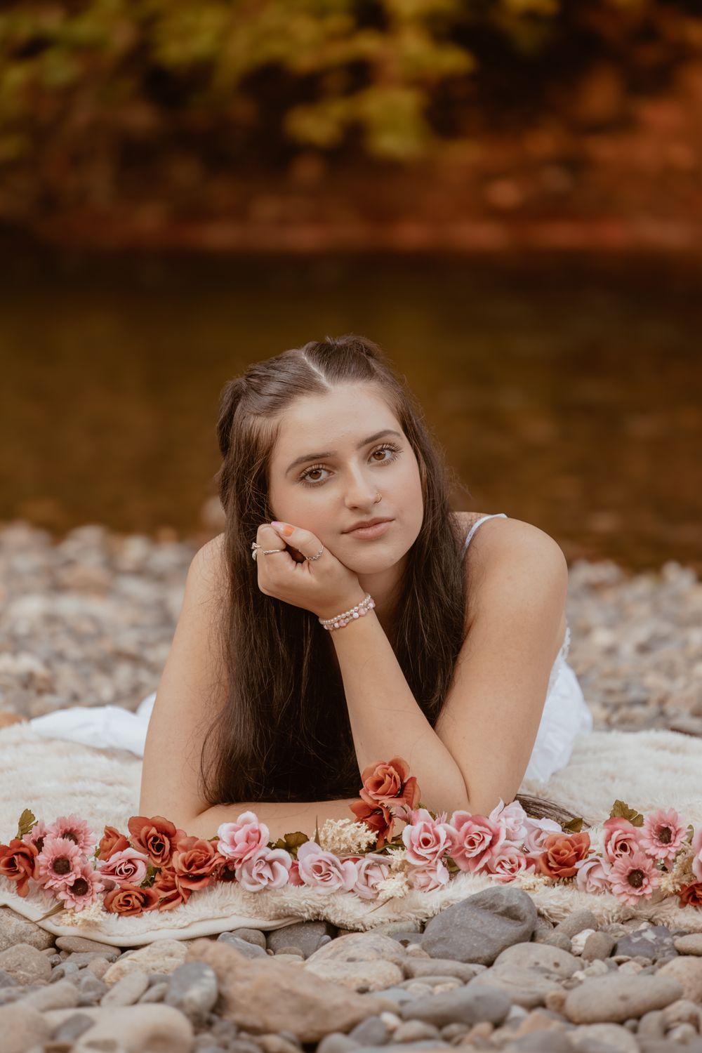 Young woman lying on rocky riverbank surrounded by pink and red flowers, wearing white dress with pearl bracelet.