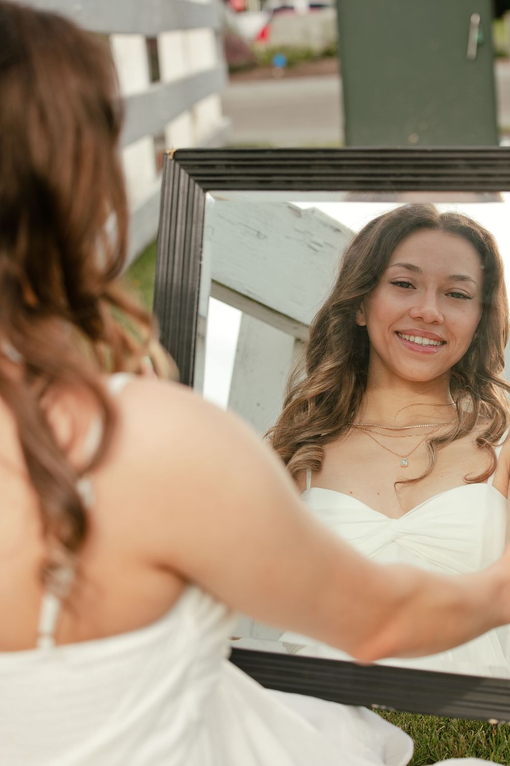 Young woman in white dress smiling at her reflection in a framed mirror outdoors.