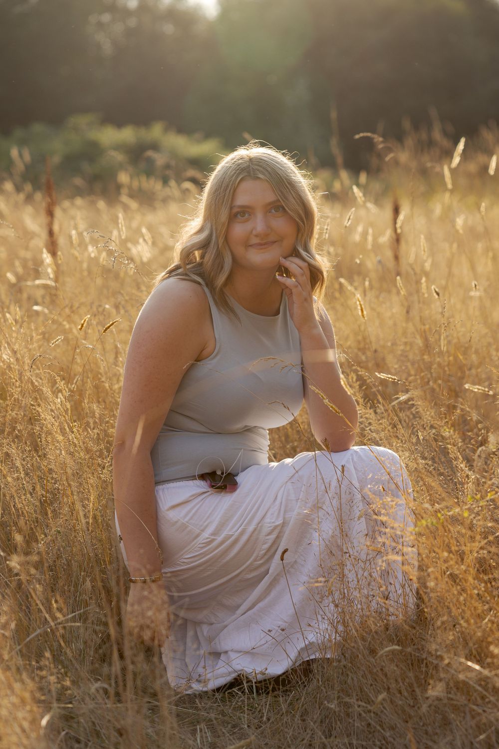 Young blonde woman in white skirt and gray top crouching in golden field at sunset.