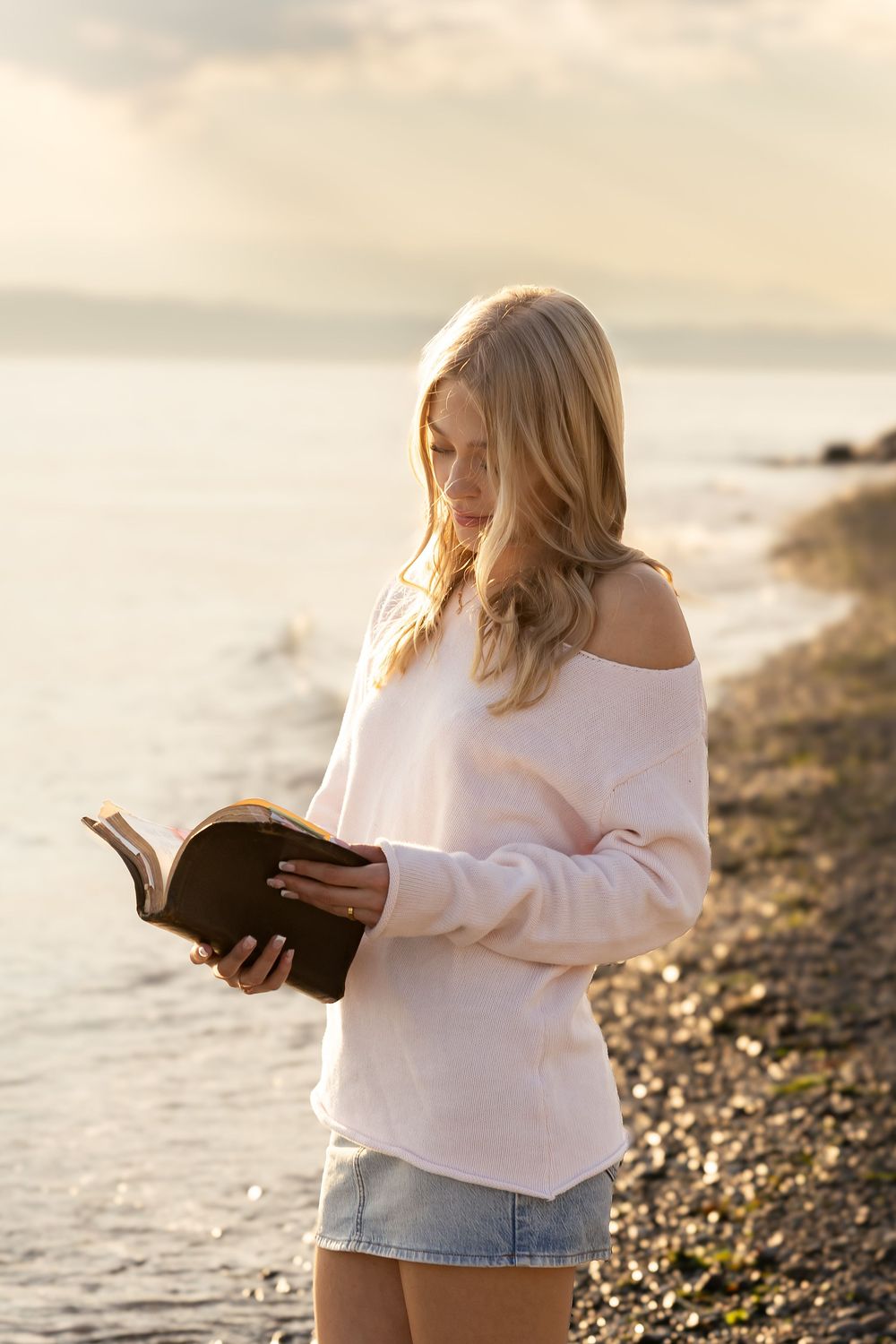 Young blonde woman reading a book on a rocky beach at sunset, wearing a white sweater and denim shorts.