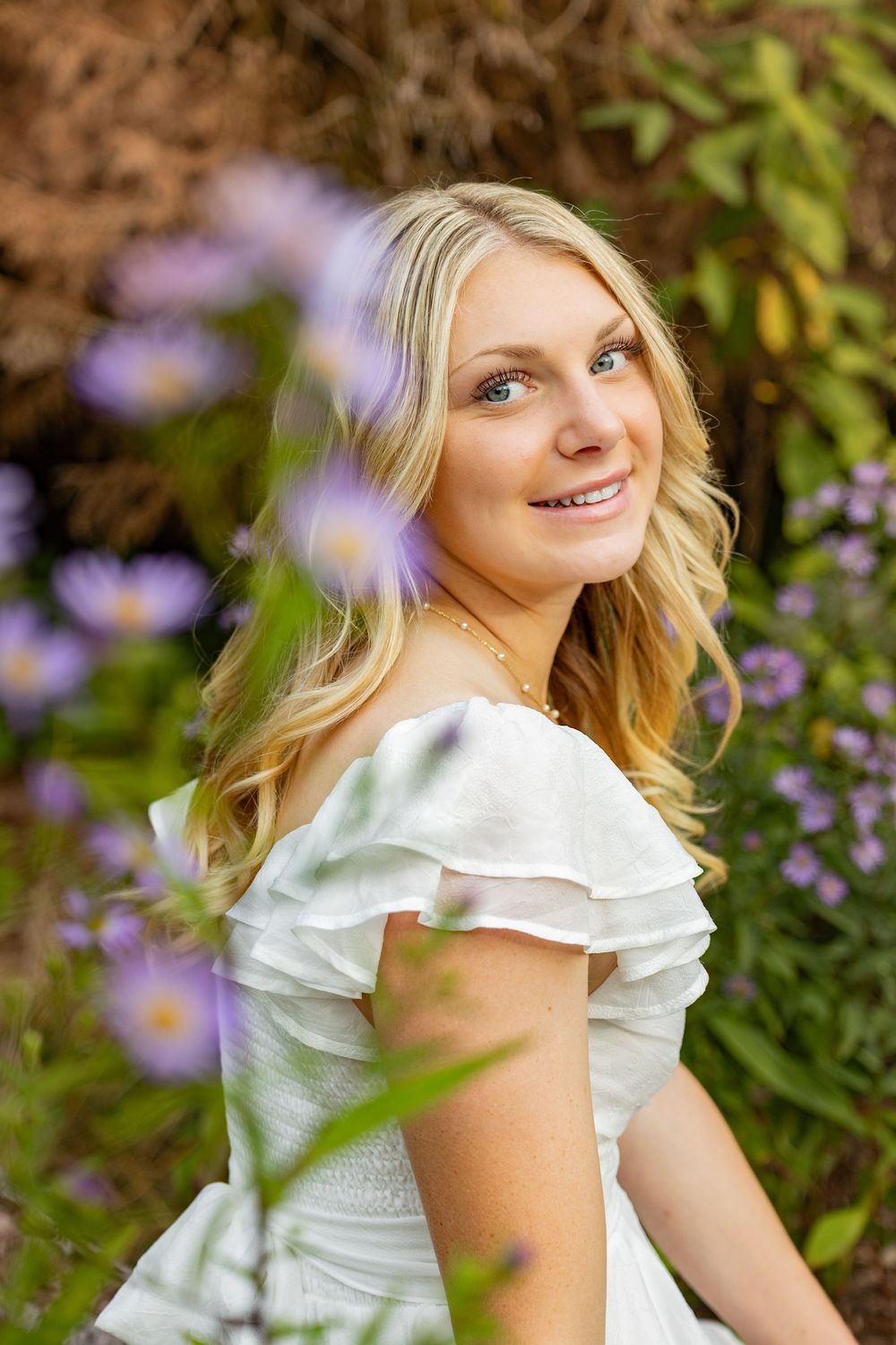 Smiling blonde woman in white ruffled dress surrounded by purple wildflowers in a lush garden setting.