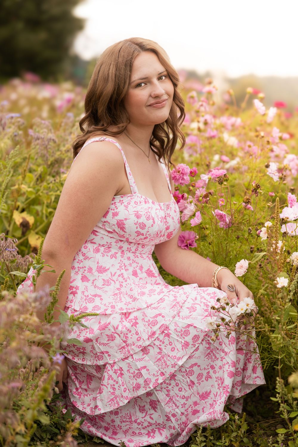 Young woman in pink floral dress sitting among wildflowers in a meadow, smiling softly at camera.