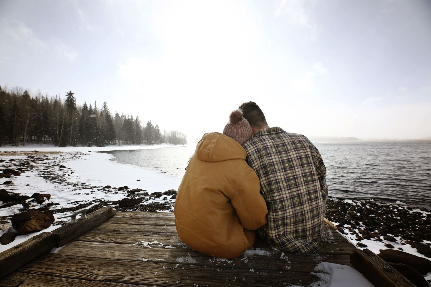 Couple in a yellow hooded jacket and plaid shirt sit close together on a wooden dock overlooking a snowy lake shoreline.