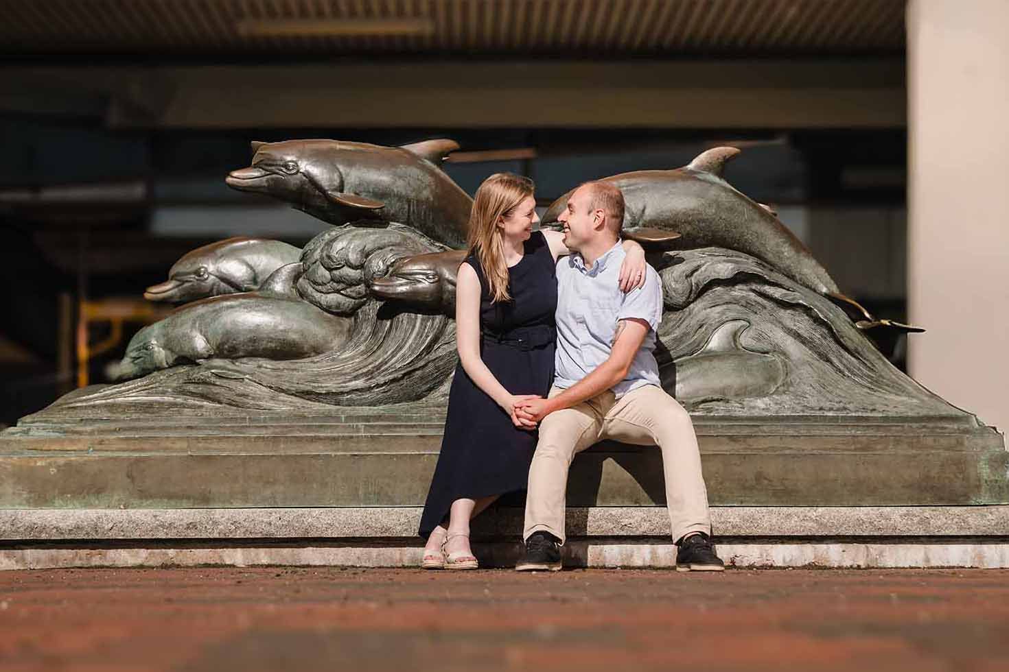 Engaged couple sitting together in front of the bronze dolphin sculpture outside the New England Aquarium in Boston