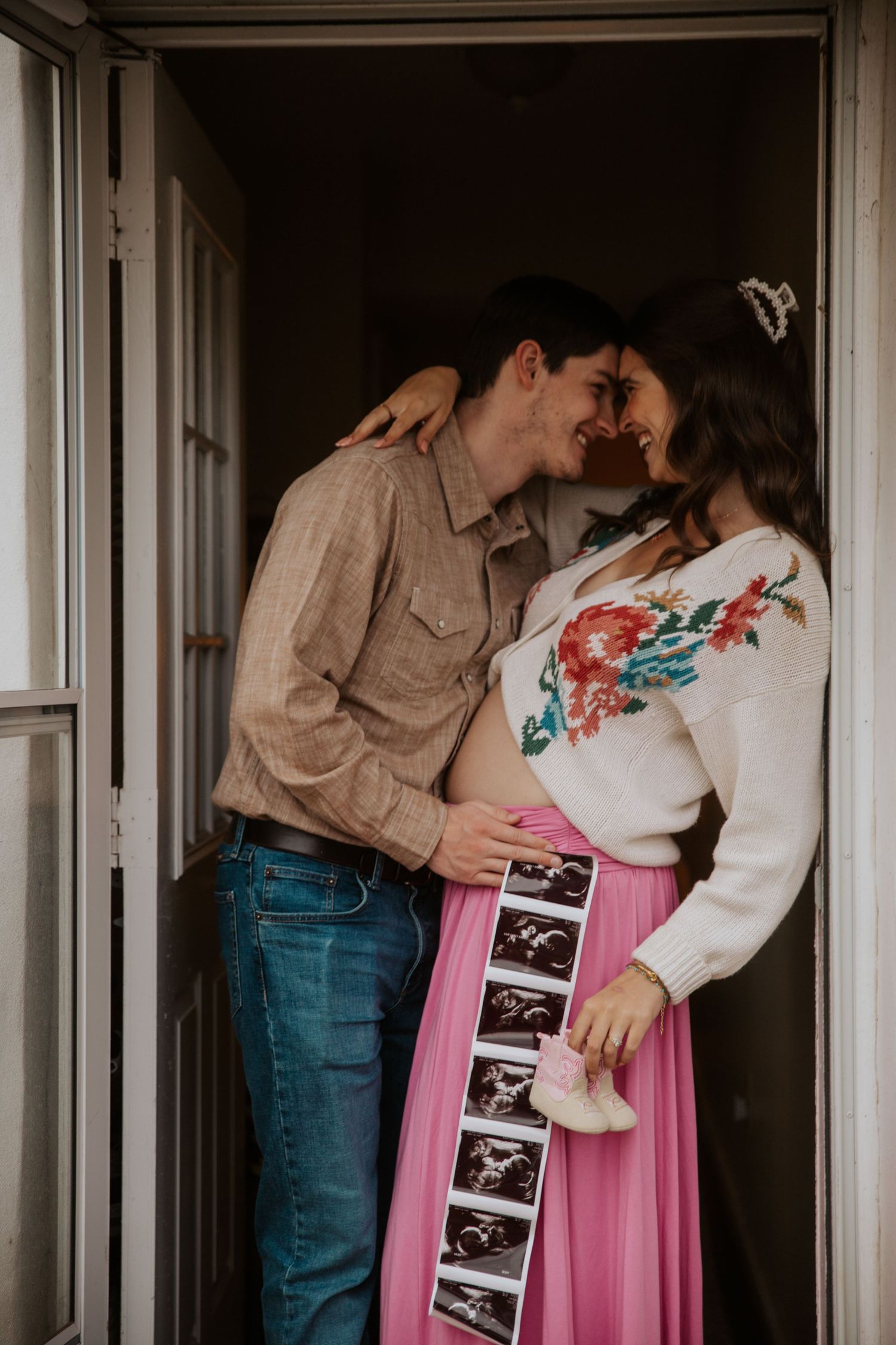 Pregnant couple sharing a tender moment in doorway, holding ultrasound photos and pink baby booties.