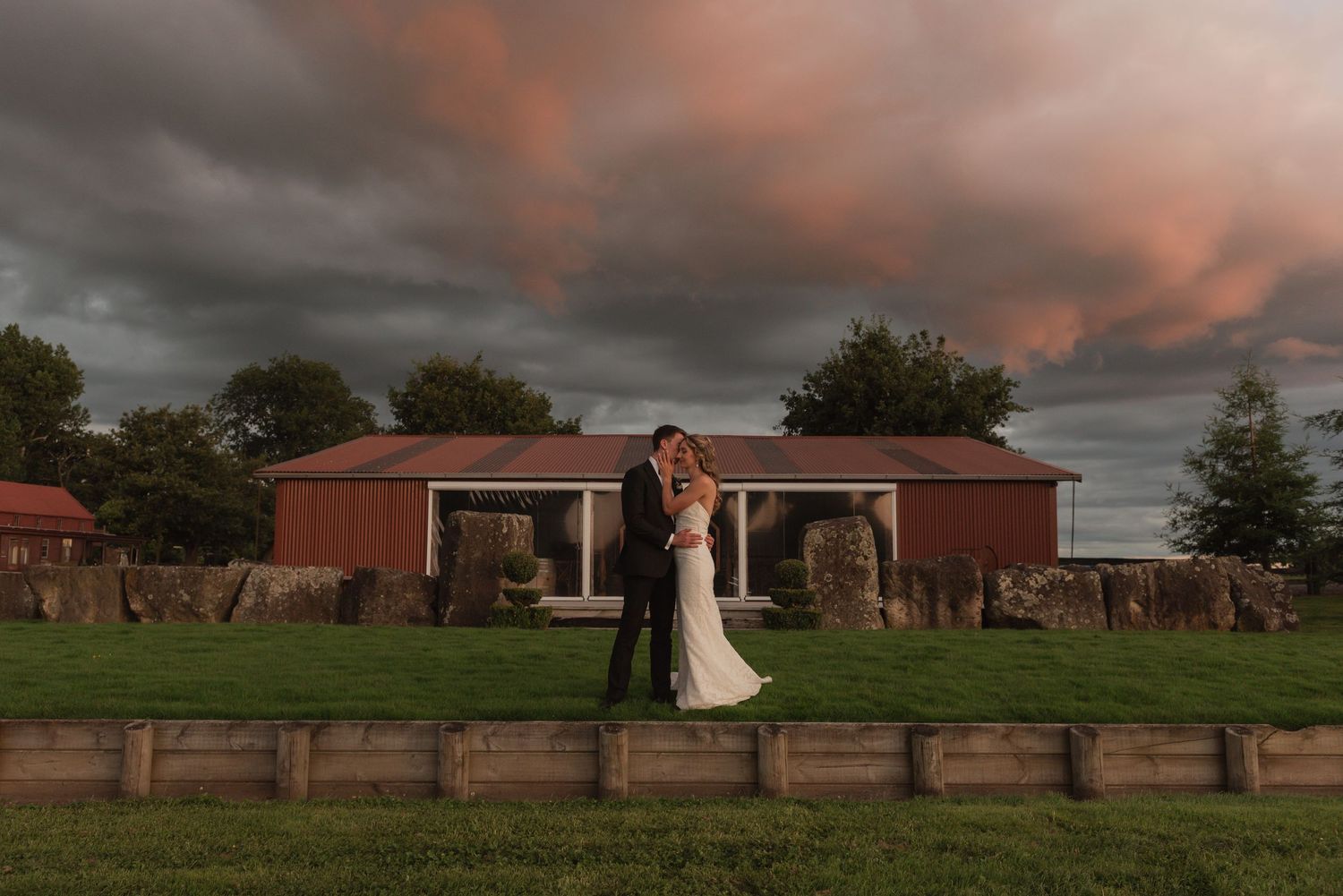 Bride and groom embrace at sunset before a red barn with dramatic storm clouds at a rustic outdoor wedding venue.