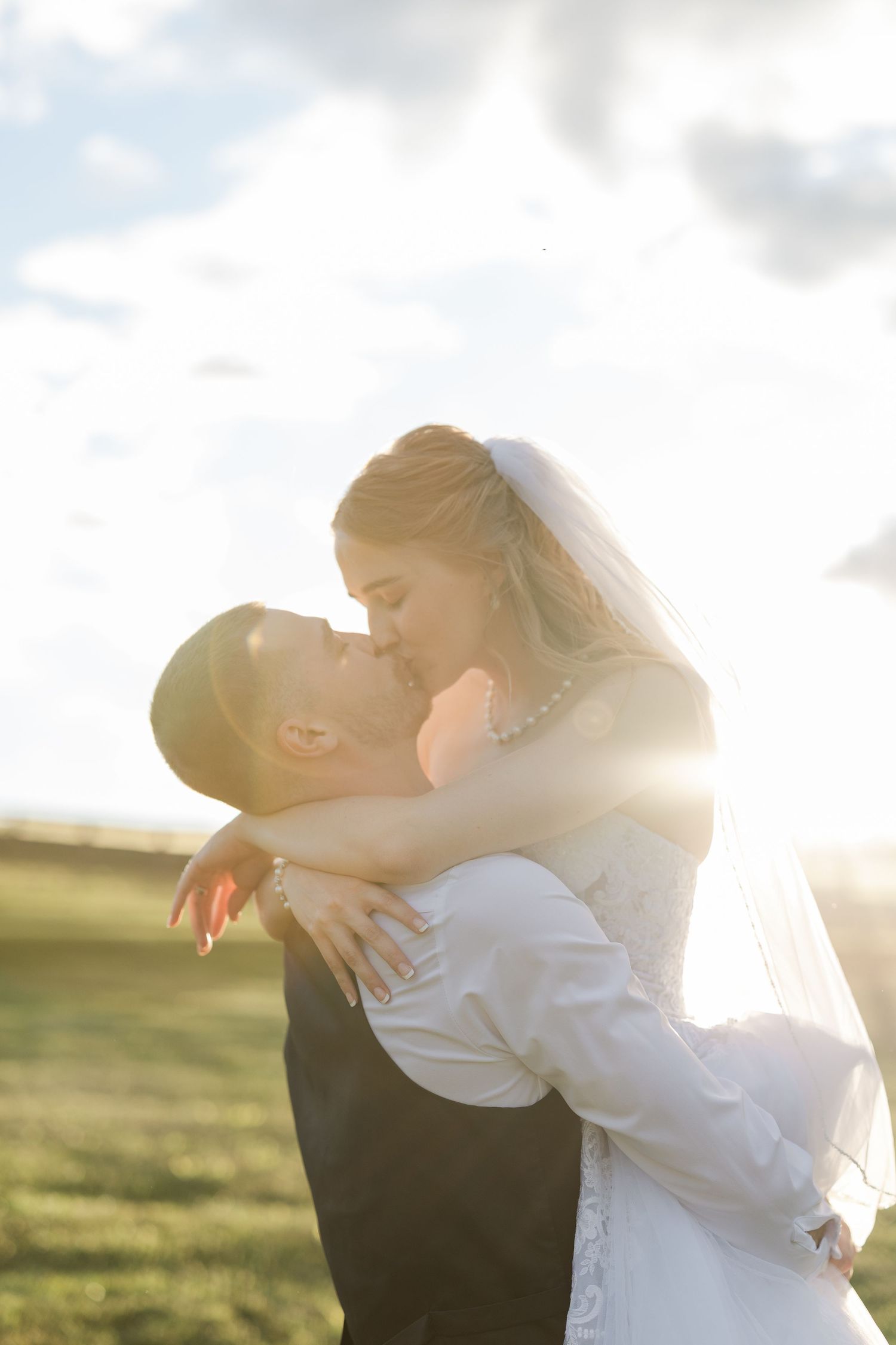 Bride and groom sharing a romantic kiss outdoors, bathed in golden sunlight with a green field and cloudy sky behind them.