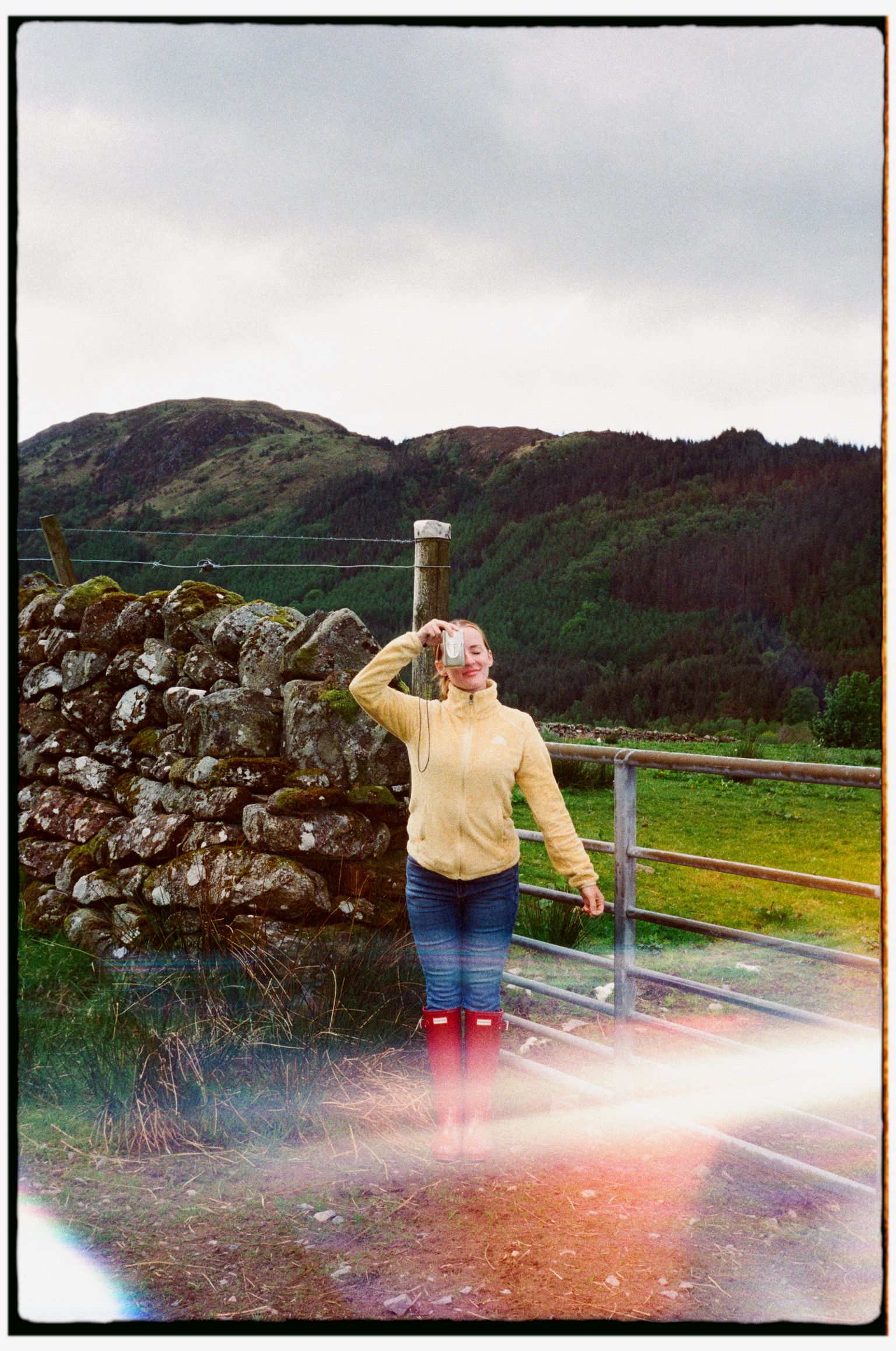 Woman in yellow sweater and red boots photographs scenery beside a stone wall with green hills in background.