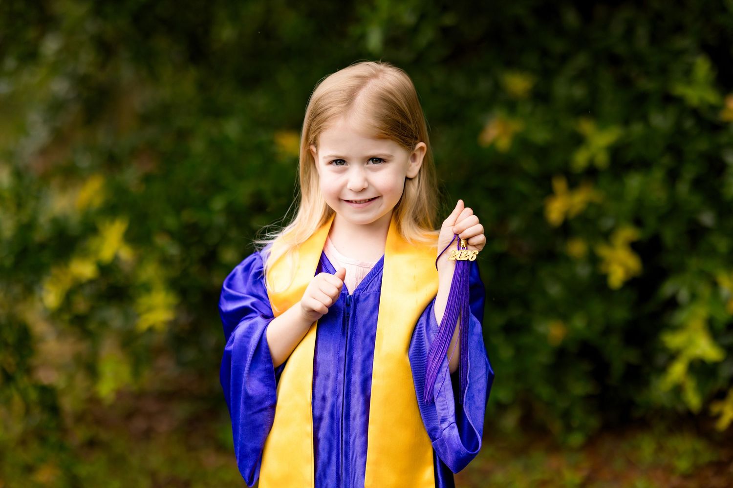 Young girl in blue and gold graduation gown smiling outdoors with green foliage background.