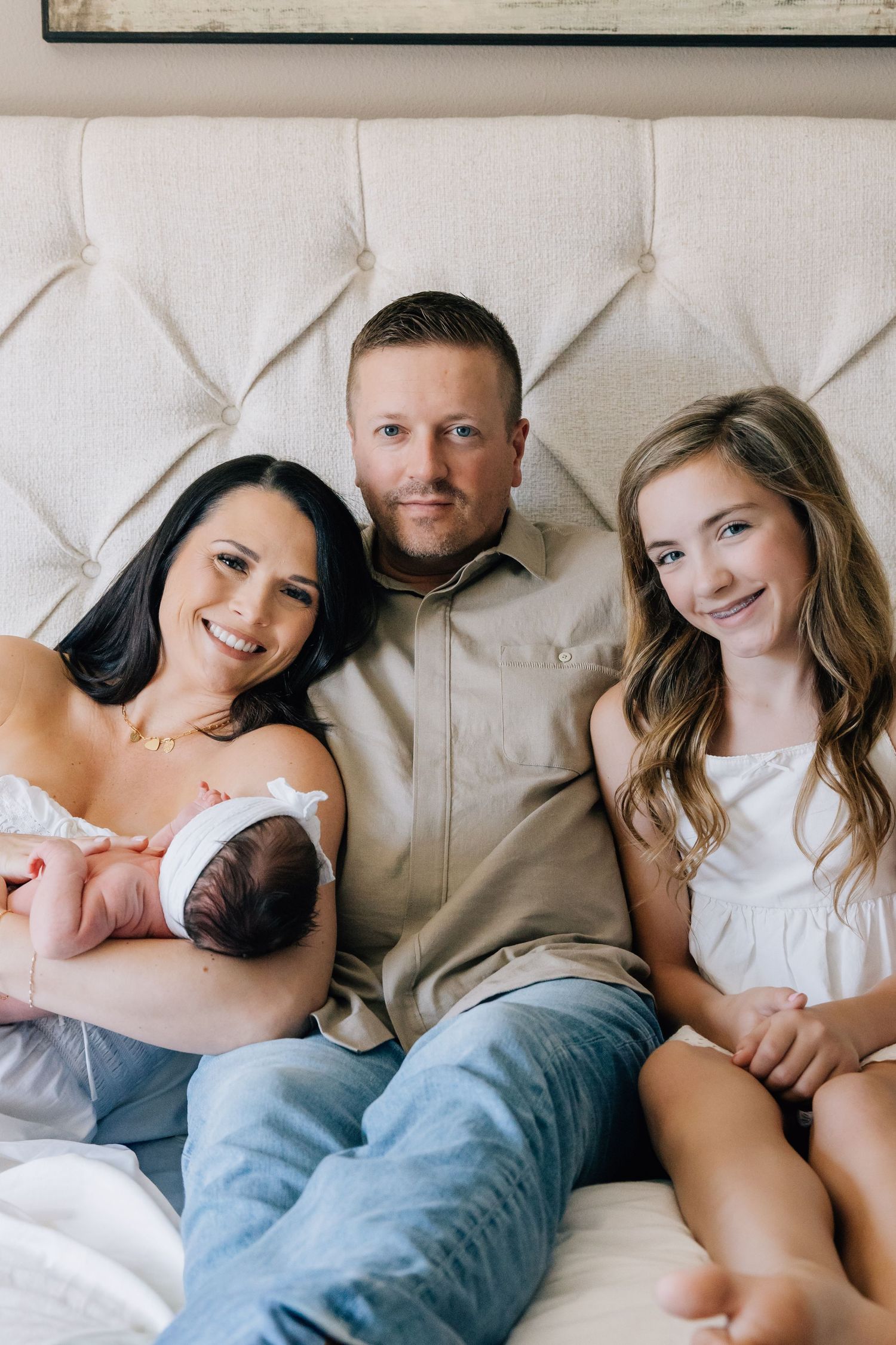Happy family of four posing on a bed, mom holding newborn baby, dad and young daughter smiling warmly.