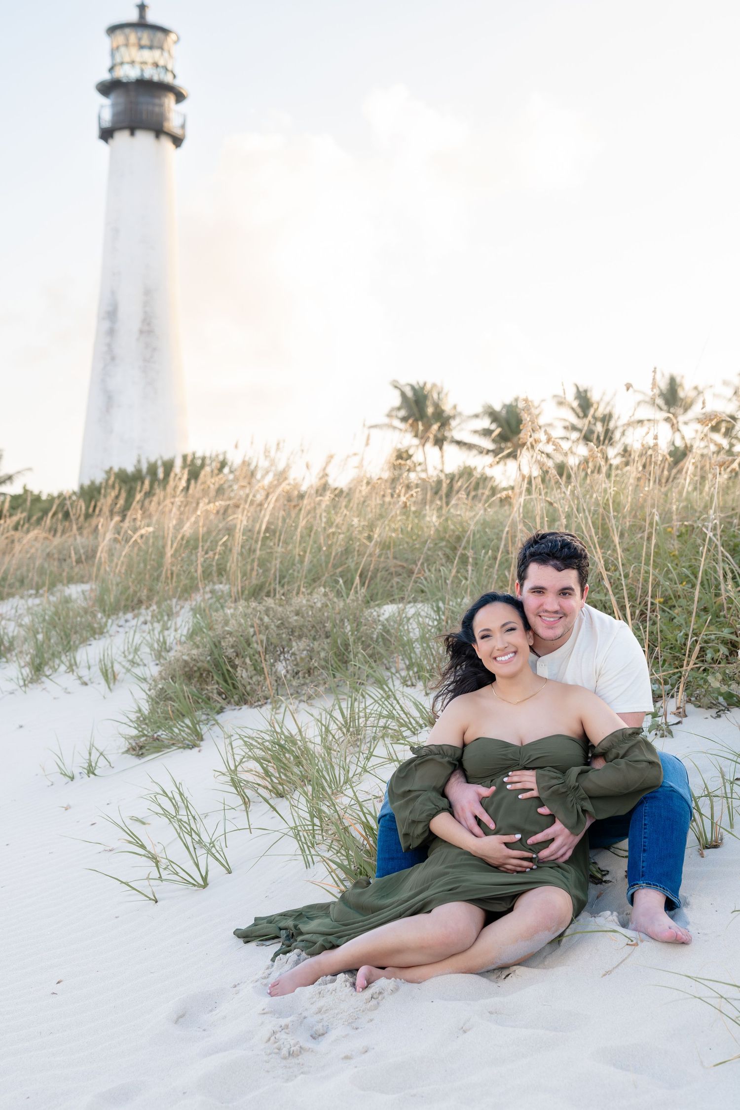 Couple sitting on white sand dunes in front of a lighthouse, woman in green maternity dress, maternity photoshoot.