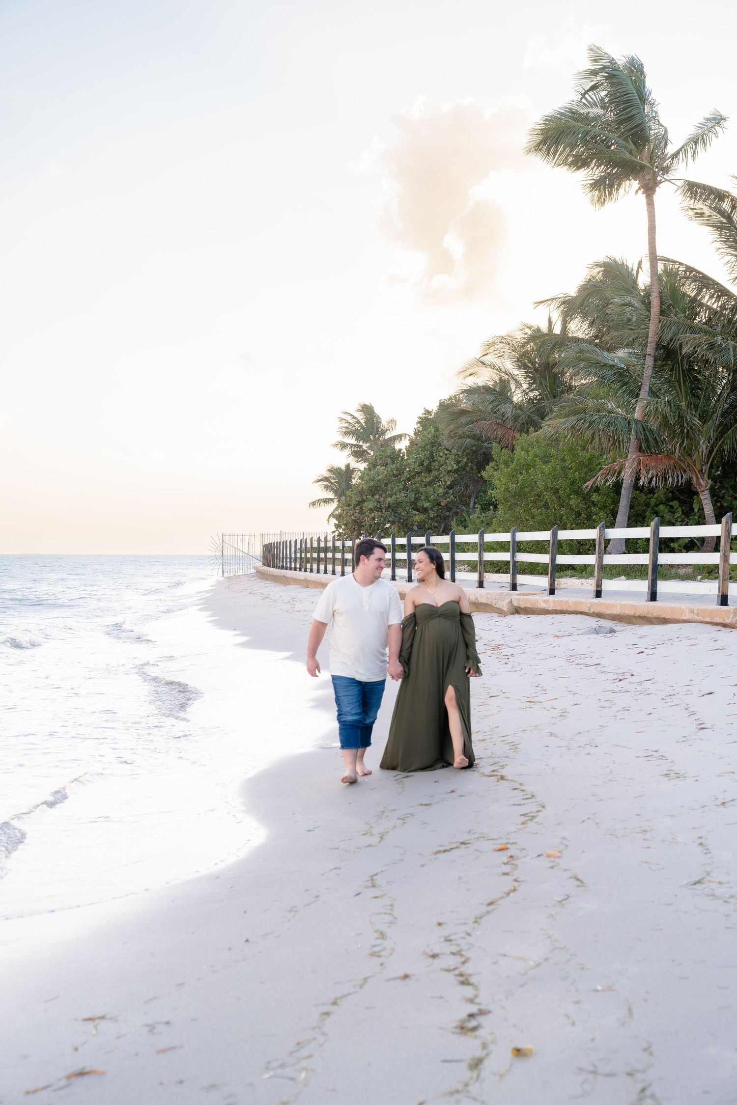 Couple walking hand-in-hand on a white sand beach at sunset, with palm trees and a white fence in the background.