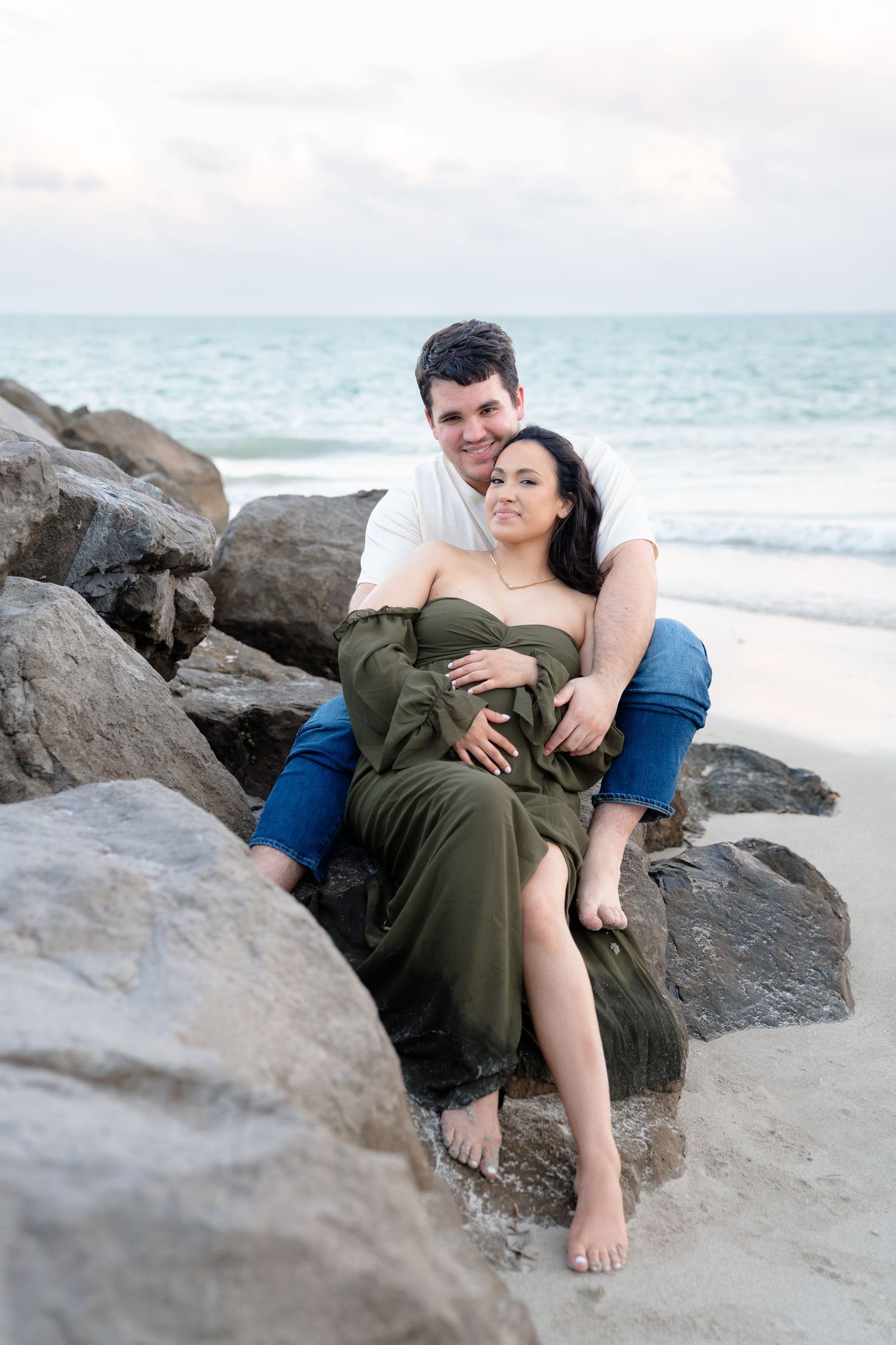 Couple sitting on beach rocks, woman in green dress, man in white shirt and jeans, ocean waves in background.