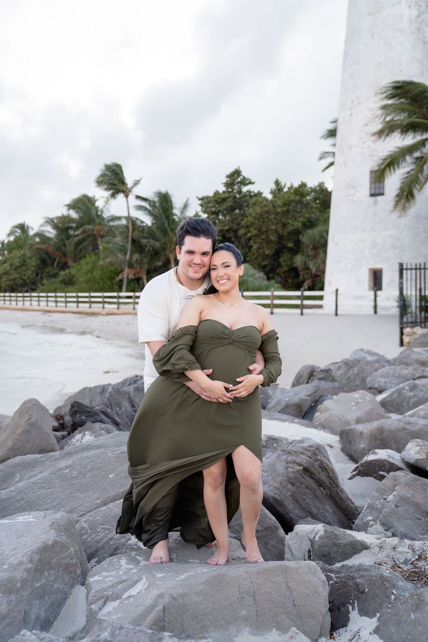 Couple poses on rocks near lighthouse, woman in olive maternity dress, man in white shirt, tropical beach backdrop.