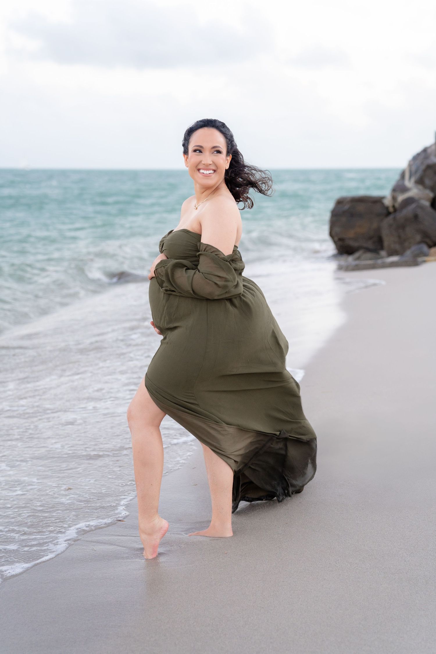 Pregnant woman in flowing olive green off-shoulder dress posing barefoot on sandy beach with ocean waves and rocks.
