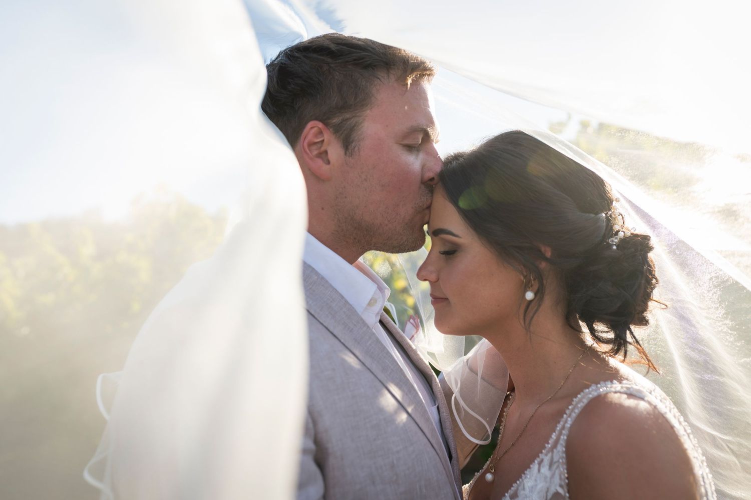 A groom kisses his bride on the forehead under a flowing veil in a romantic outdoor wedding portrait.