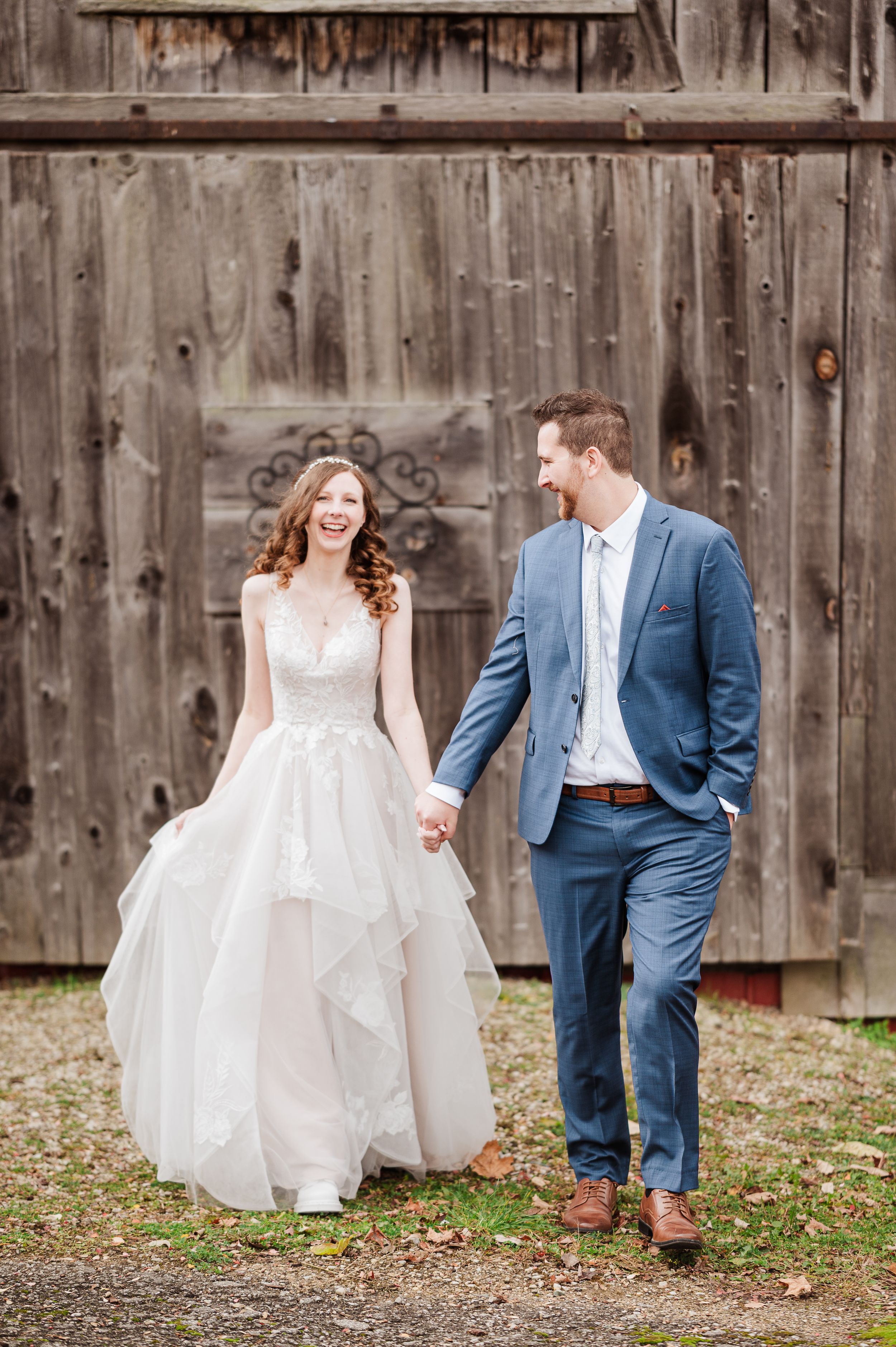 A joyful bride in a white gown and groom in a blue suit hold hands walking before a rustic barn.
