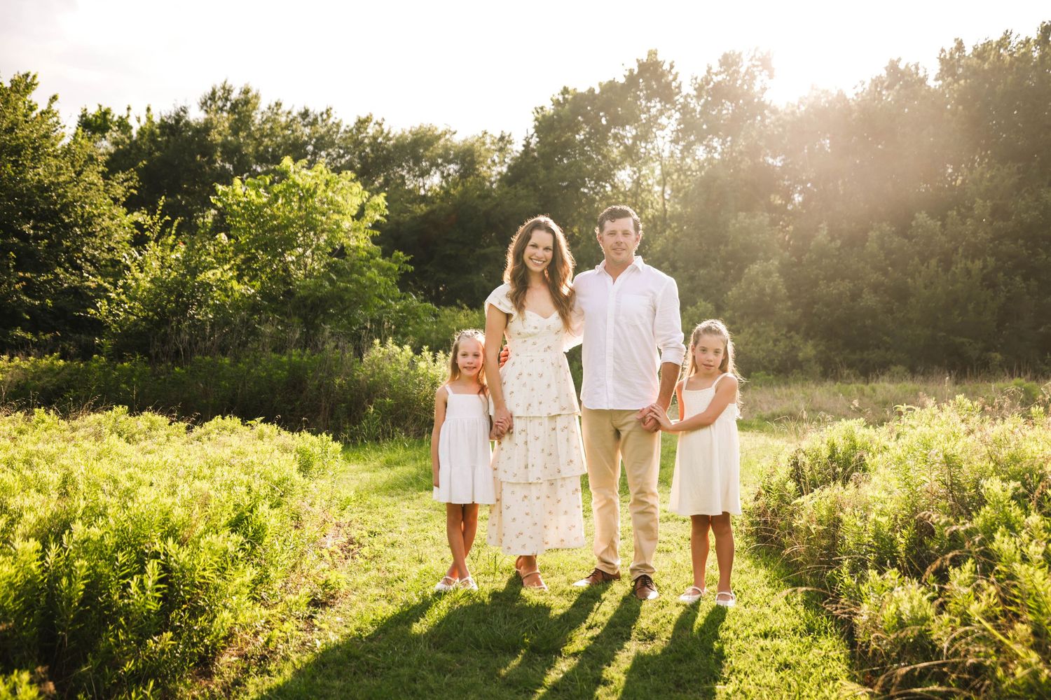 A happy family of four walks through a sunny green meadow, parents holding hands with two girls in white dresses.