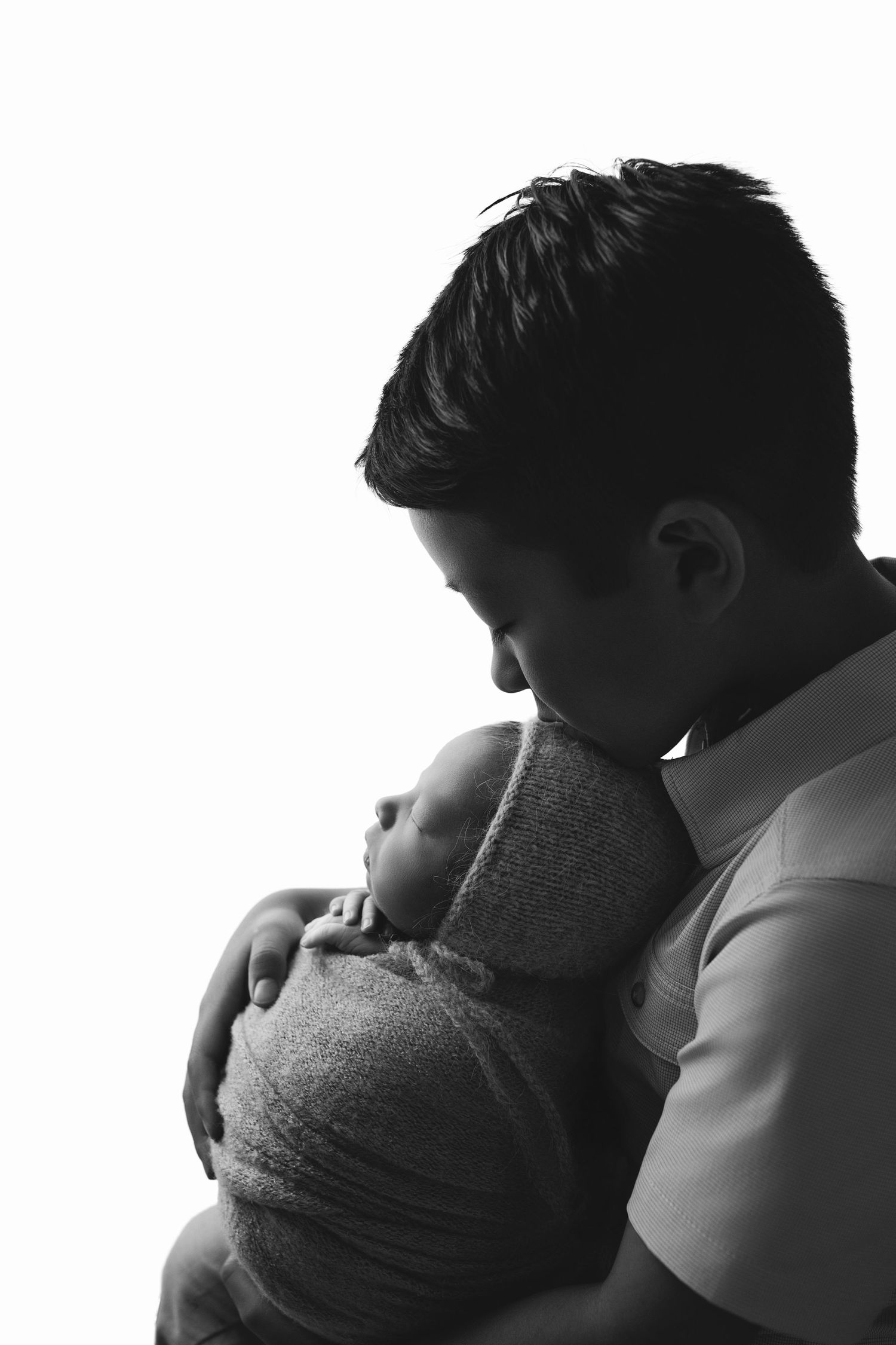 Black and white photo of a father tenderly kissing his sleeping newborn baby wrapped in a gray swaddle.