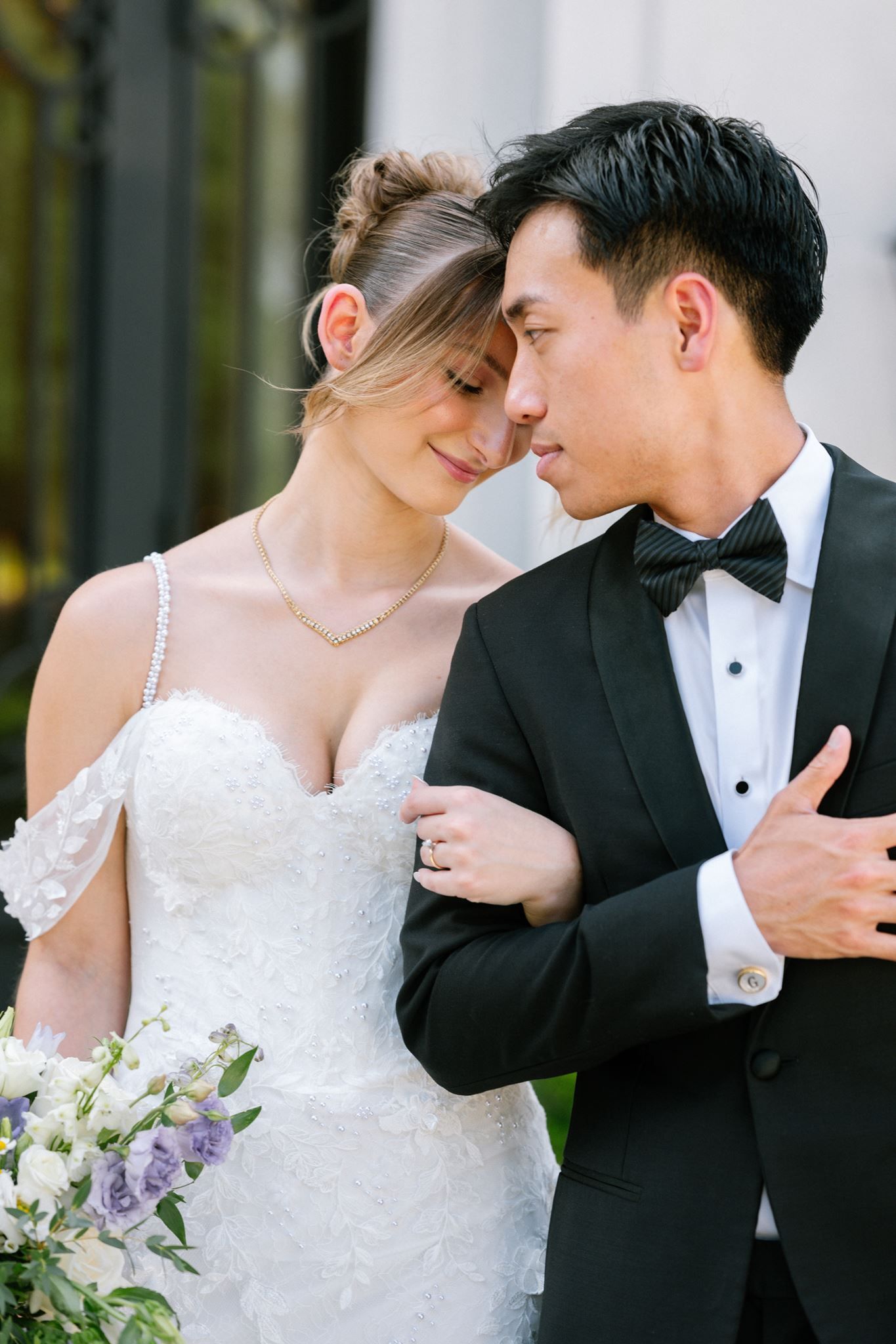 A bride in a white lace gown and groom in a black tuxedo share an intimate moment, foreheads touching, holding a floral bouquet.