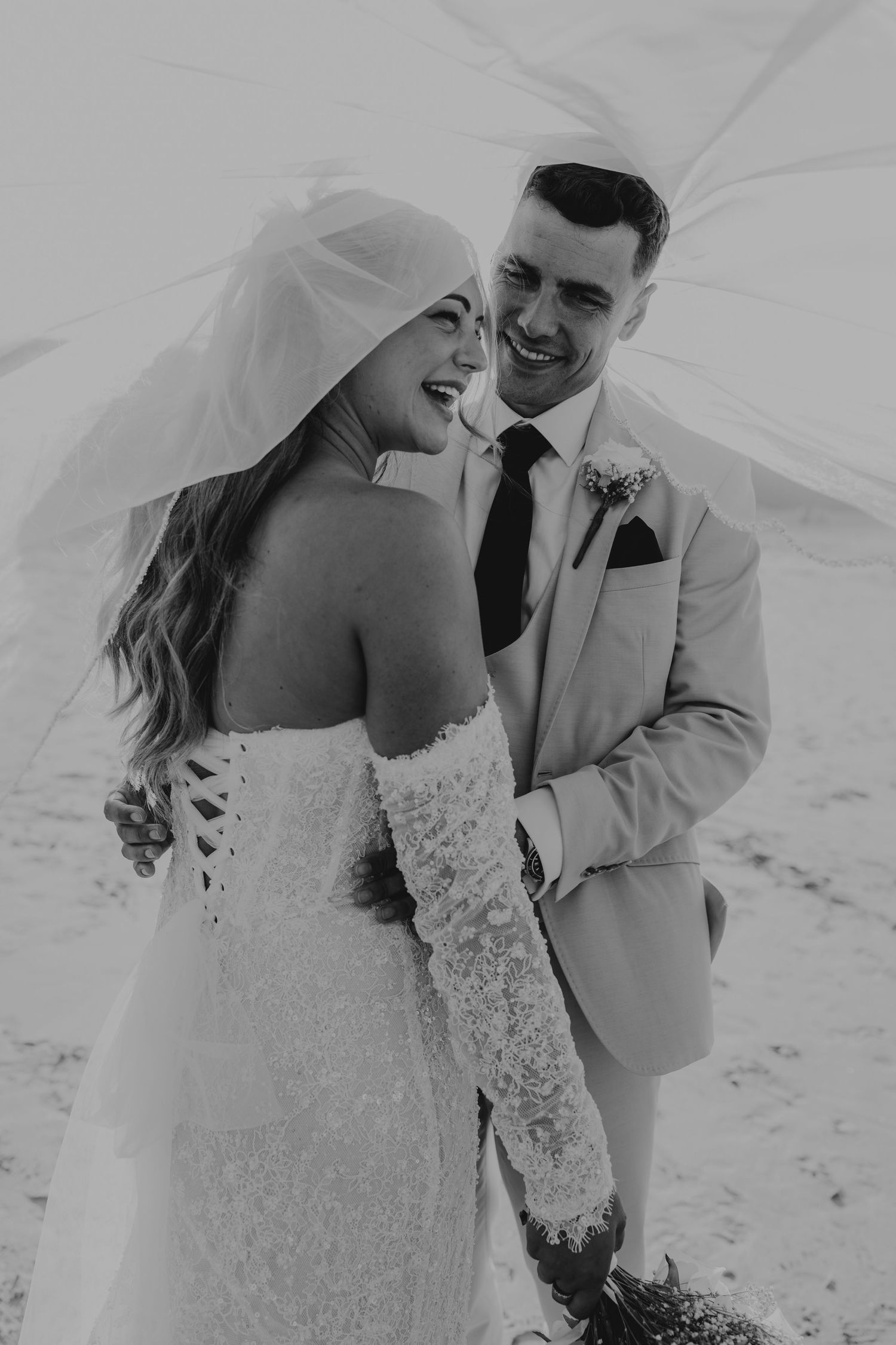 Black and white photo of a smiling bride and groom embracing under a flowing veil on their wedding day.