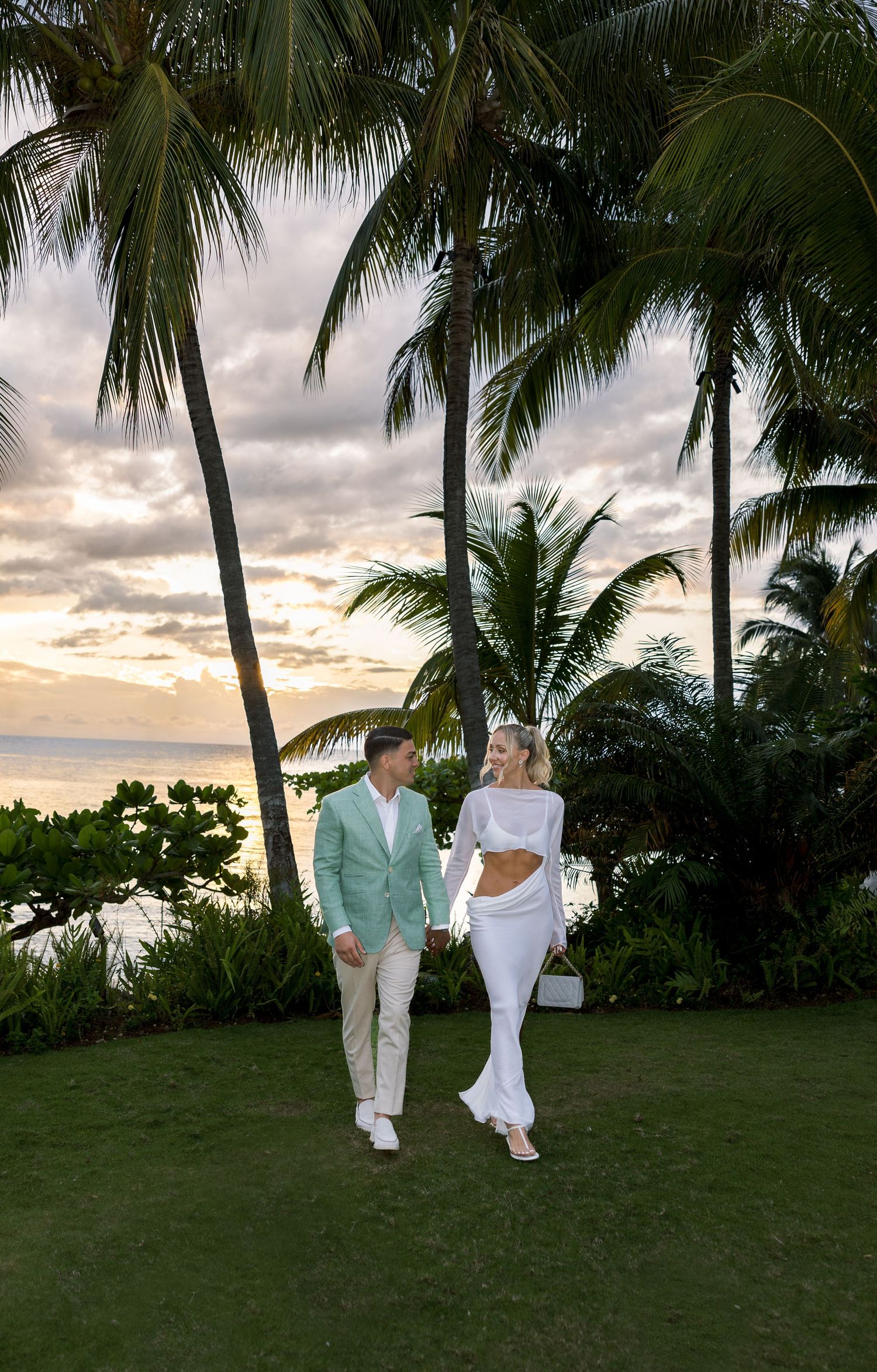 Bride and Groom stand by a tropical resort pool with tall palm trees and ocean views under a clear blue sky at Round Hill Hotel & Villas