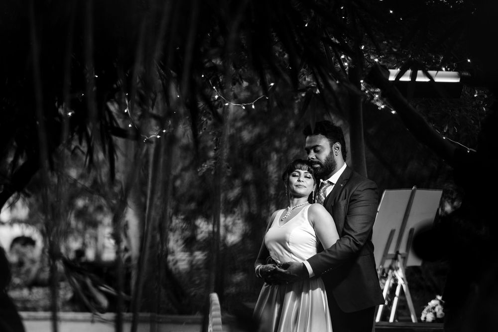 Black and white photo of a couple embracing outdoors at a romantic evening wedding ceremony under trees with string lights.
