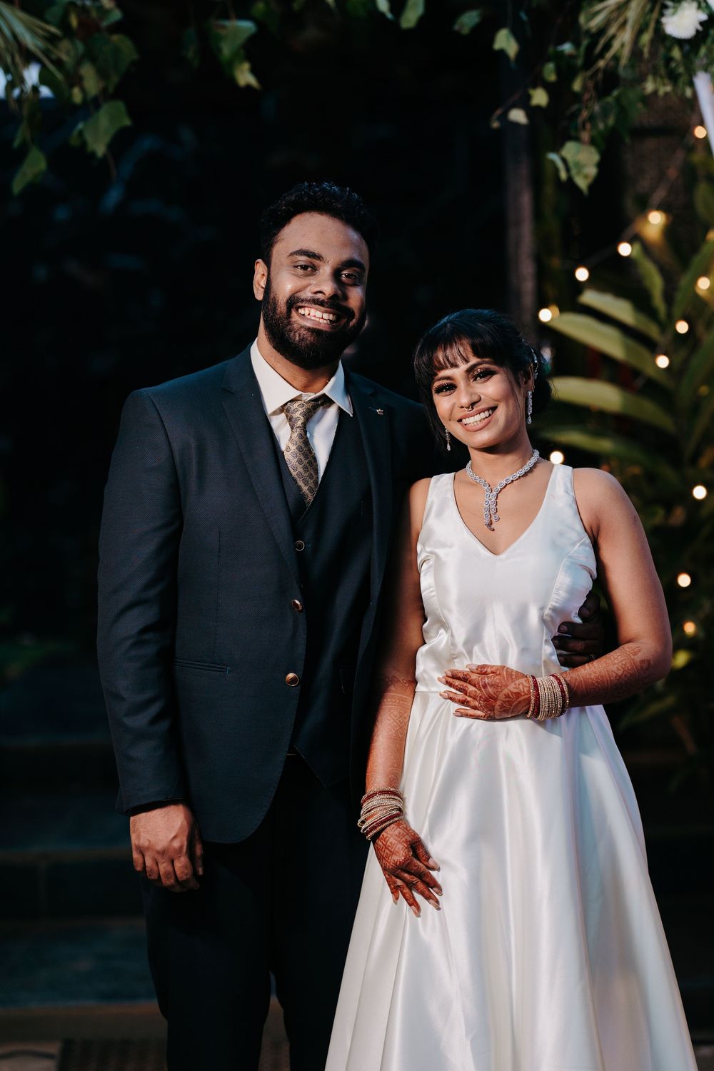 Smiling bride in white gown and groom in dark suit pose together at an elegant outdoor wedding reception.