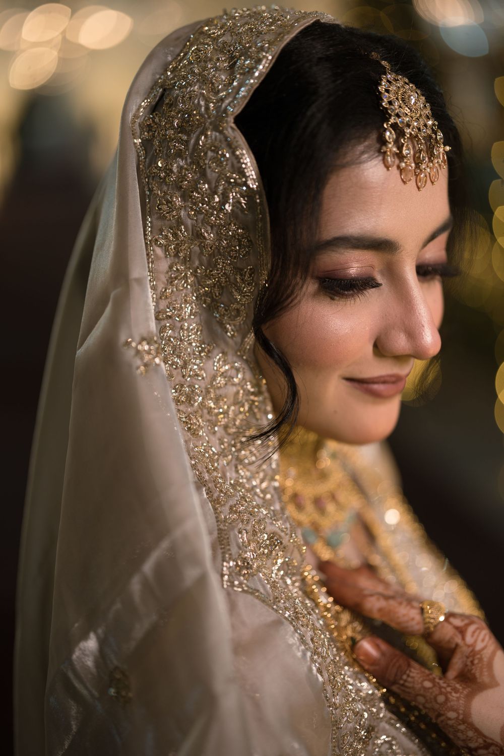 A bride in gold-embroidered white dupatta and maang tikka looks down gracefully, with henna hands and glowing makeup.