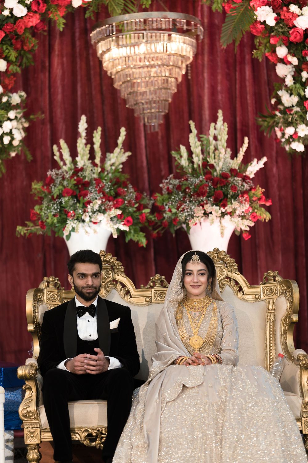 South Asian bride and groom in traditional wedding attire seated on ornate gold chairs beneath a crystal chandelier.