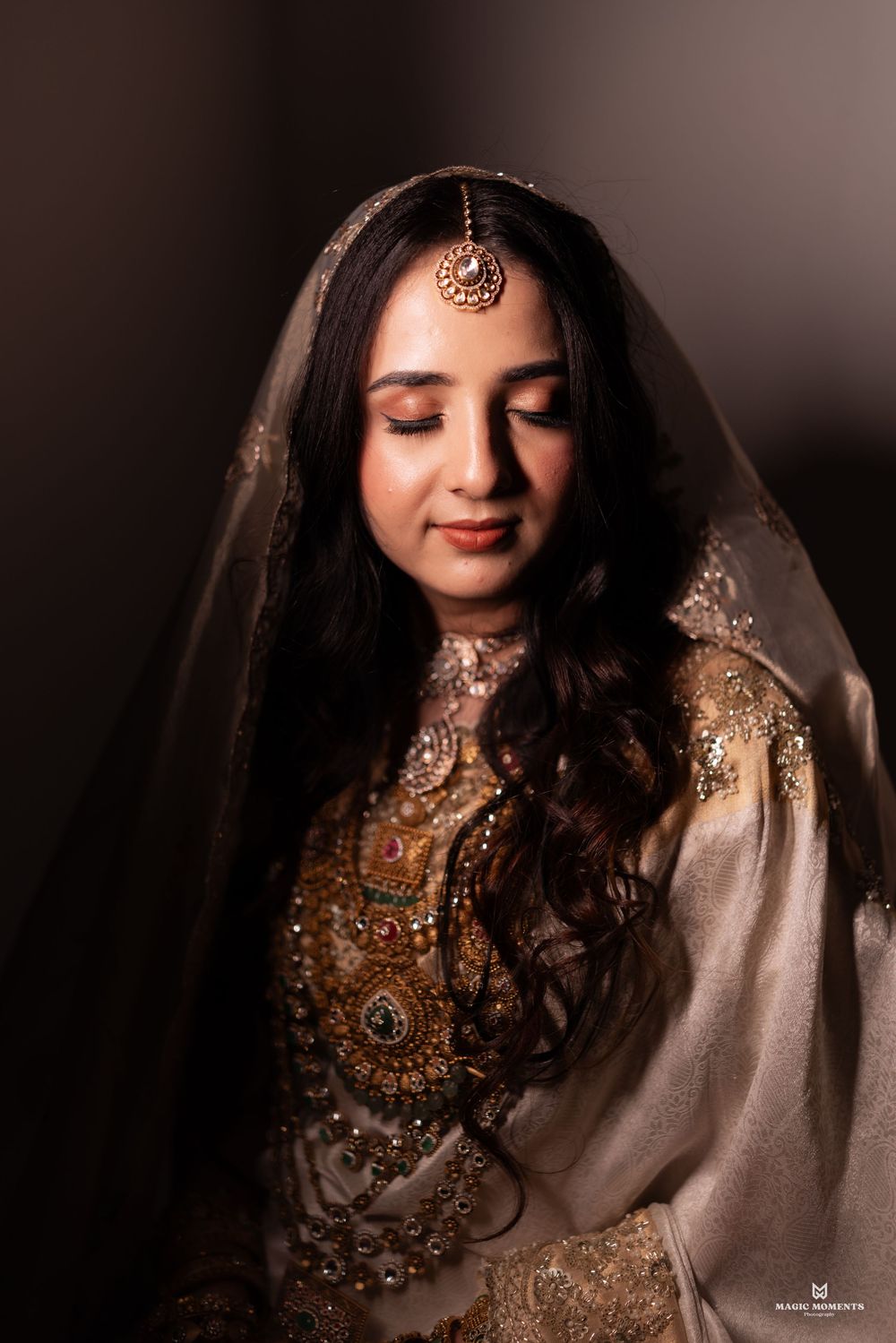 A bride in elegant gold embroidered attire with a veil and maang tikka, eyes closed in a serene pose.
