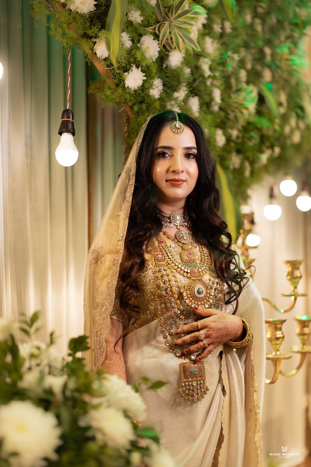 Bride in white saree with gold jewelry and dupatta poses at elegant floral wedding decor backdrop.