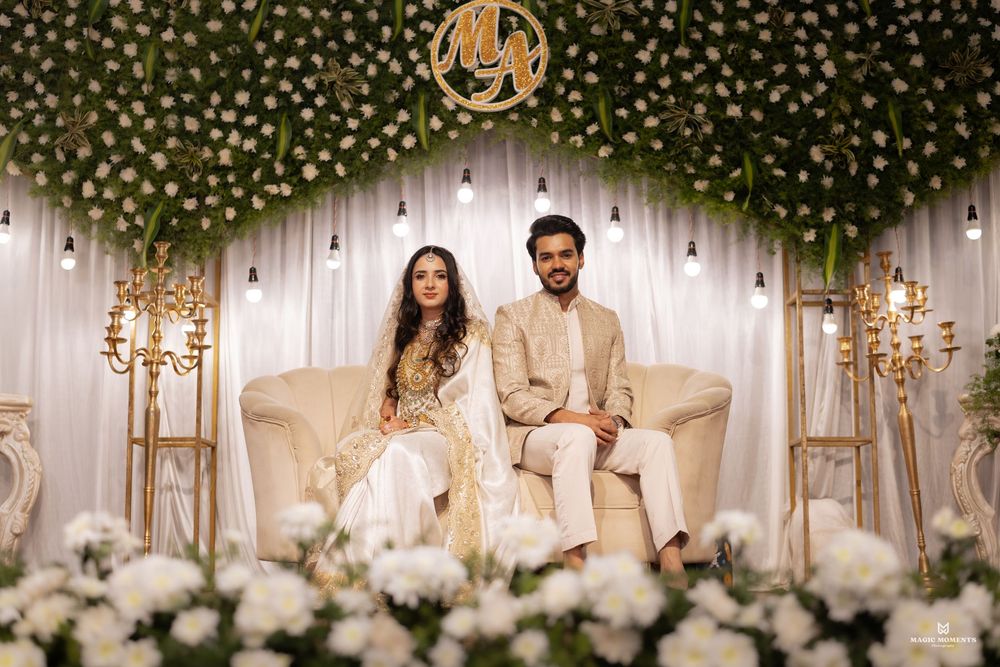 Elegant couple seated on white sofa at floral wedding backdrop with greenery arch and gold monogram decor.