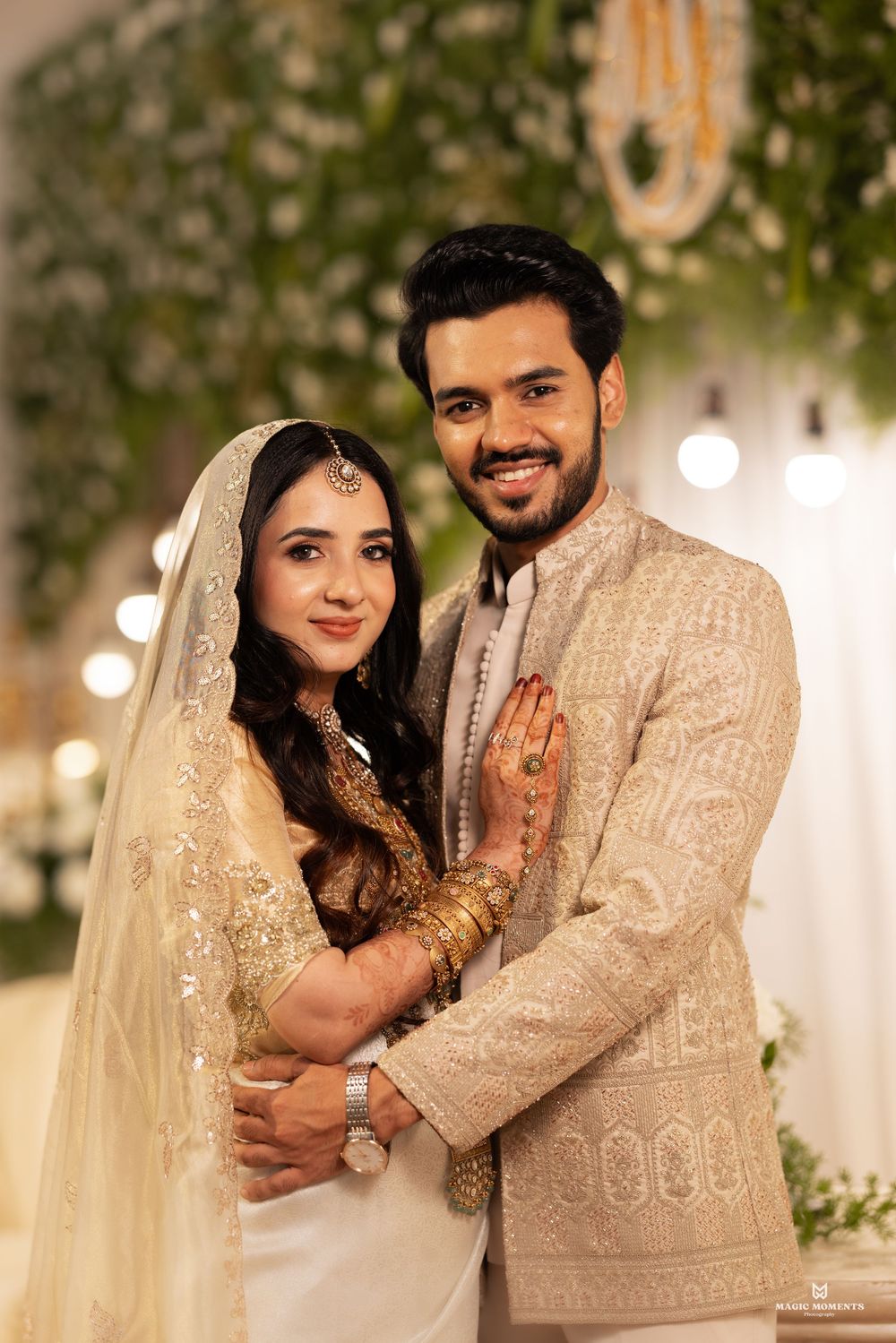 Elegant South Asian couple in ivory wedding attire posing together at a decorated ceremony venue.