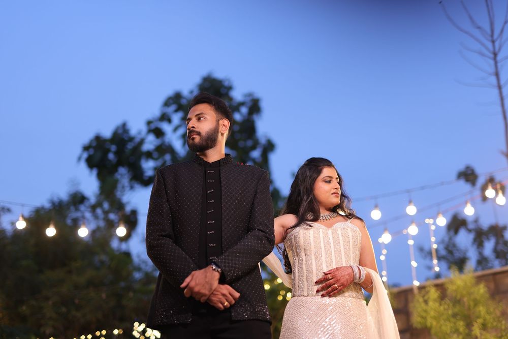 Couple in elegant Indian attire posing outdoors at dusk with string lights and trees in the background.
