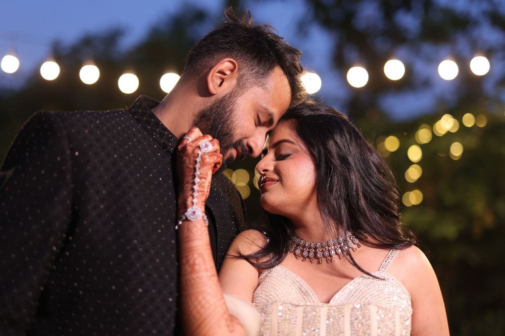 A romantic South Asian couple shares an intimate moment under string lights at dusk, the woman in white and man in black.