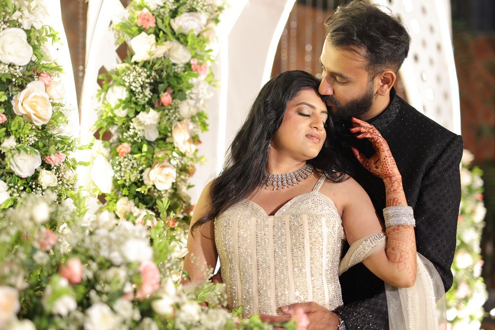 South Asian couple in wedding attire embrace near floral arch with roses, bride wearing mehndi and silver jewelry.